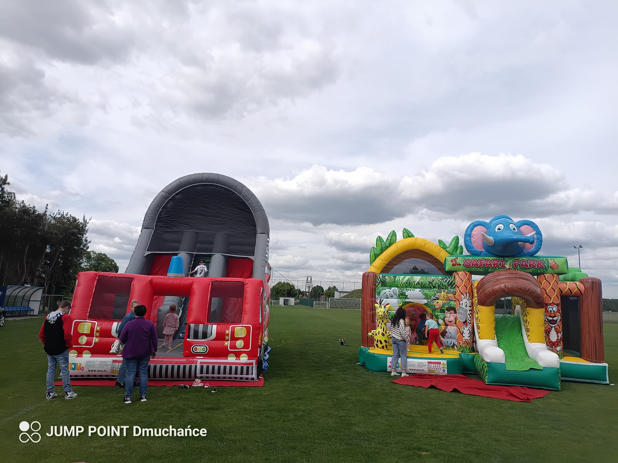 Dwa dmuchańce: czerwony w kształcie wozu strażackiego z tunelem i Safari Park ze zjeżdżalnią. Dzieci wchodzą do środka. Trawa, pochmurne niebo w tle.