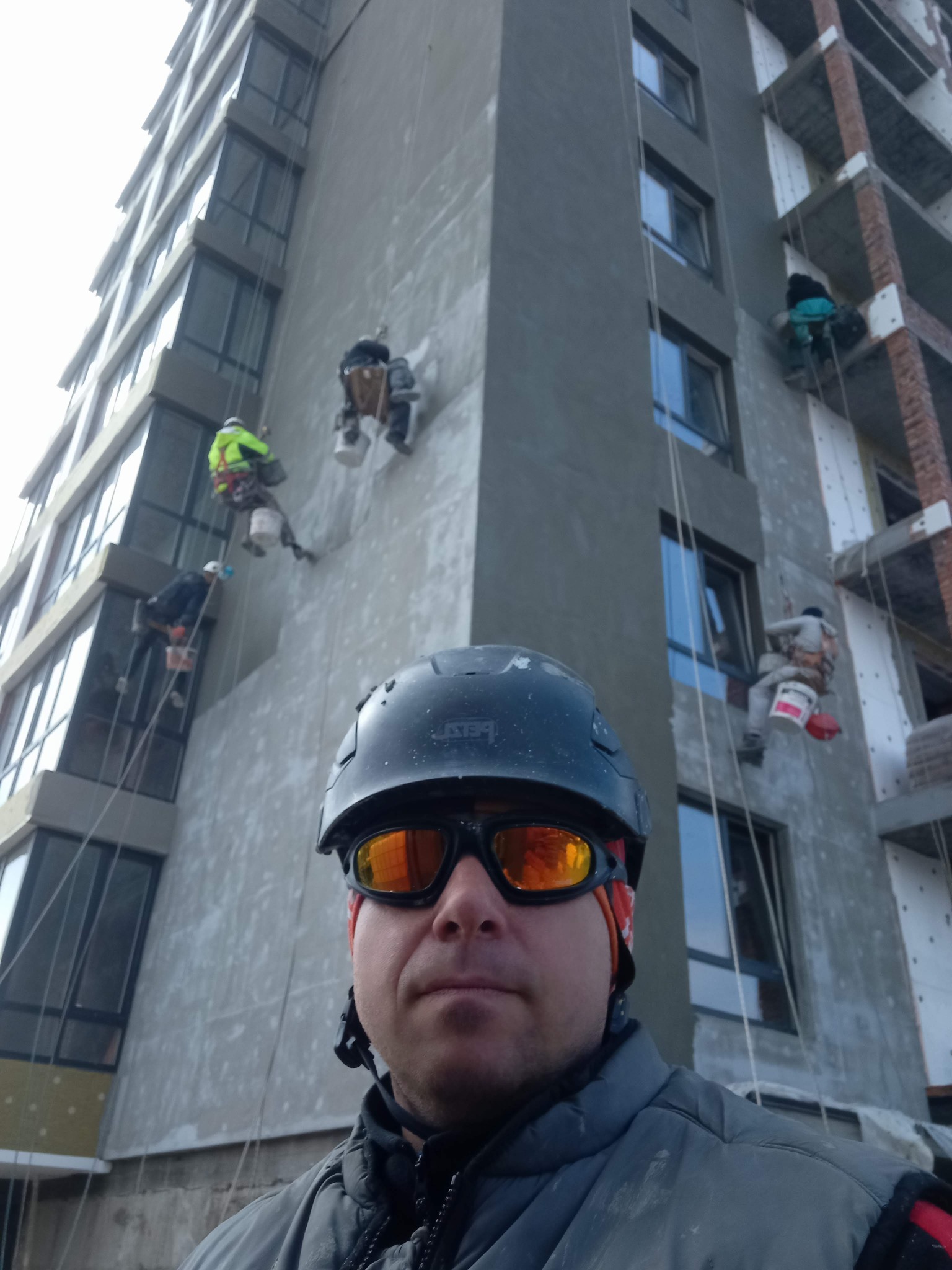 Pracownicy na linach, w kaskach i okularach, wykonują prace na elewacji wysokiego budynku. Widoczne liny, wiadra i fragment ocieplenia. Ujęcie z dołu, z bliska na jednego z pracowników.