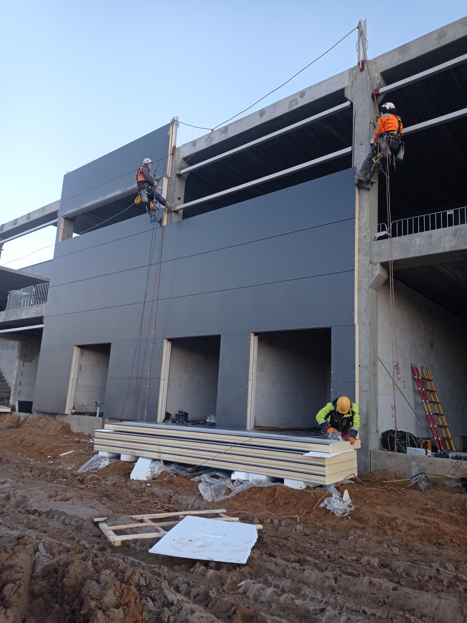 Montaż paneli elewacyjnych na wysokości przez dwóch pracowników z użyciem lin. Trzeci pracownik obrabia panele na ziemi. Budynek w stanie surowym.