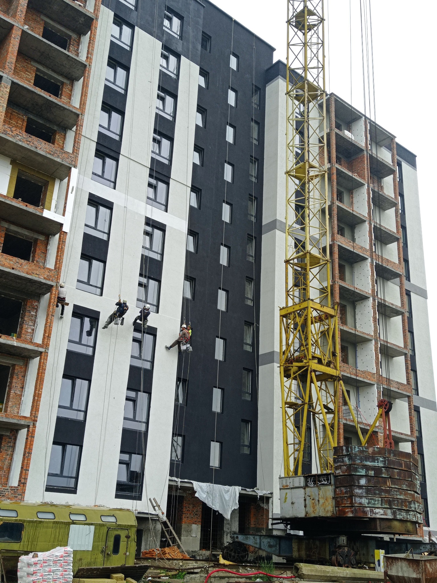 Prace wysokościowe: czterech alpinistów maluje elewację bloku w trakcie budowy. Żółty dźwig budowlany w tle. Widok z dołu na budynek.