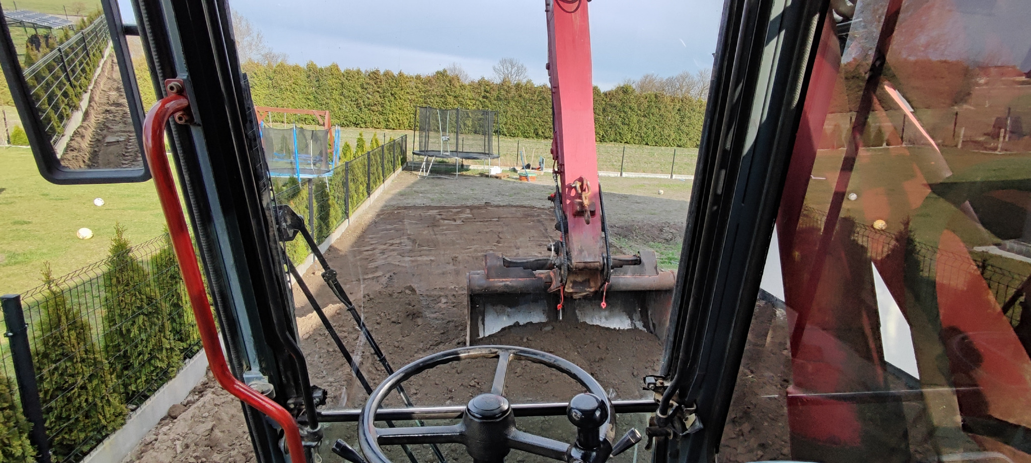 Widok z kabiny koparki na wyrównywany teren działki z trampolinami i ogrodzeniem. W tle zielone tuje. Praca w toku, łyżka koparki wypełniona ziemią.