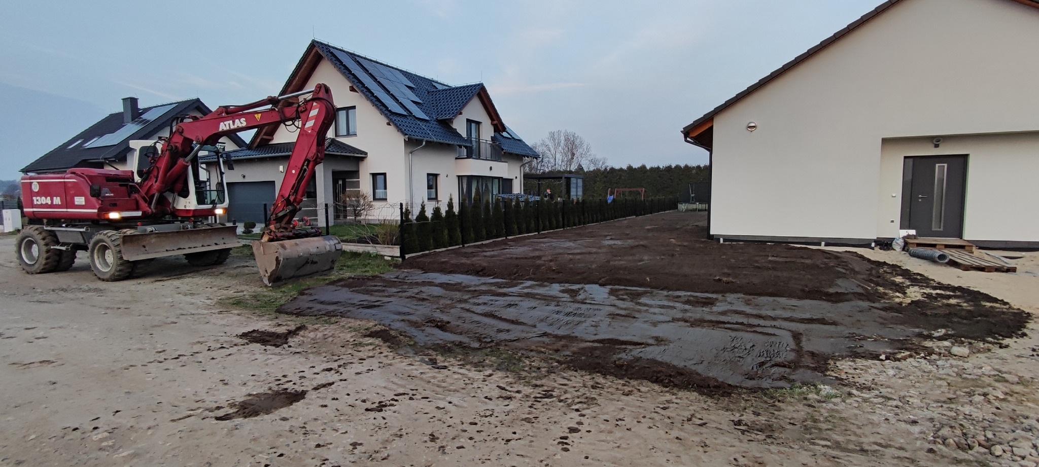 Czerwona koparka Atlas 1304 M na placu budowy, wyrównująca teren pod trawnik przed nowoczesnym domem z panelami słonecznymi. Widoczne świeże ślady opon.