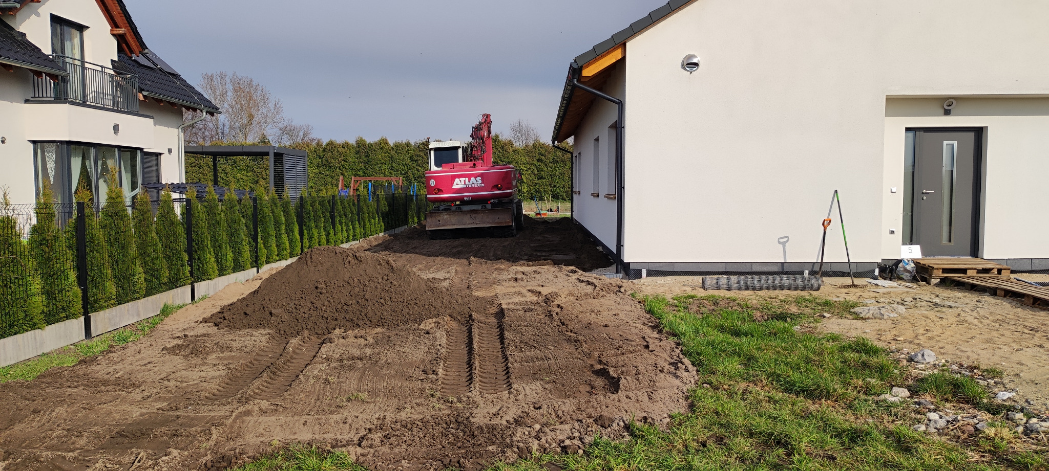 Czerwona koparka Atlas na działce, przygotowuje teren pod budowę. Ślady opon na ziemi, w tle domy i zielone tuje. Dzień słoneczny, widoczne narzędzia.