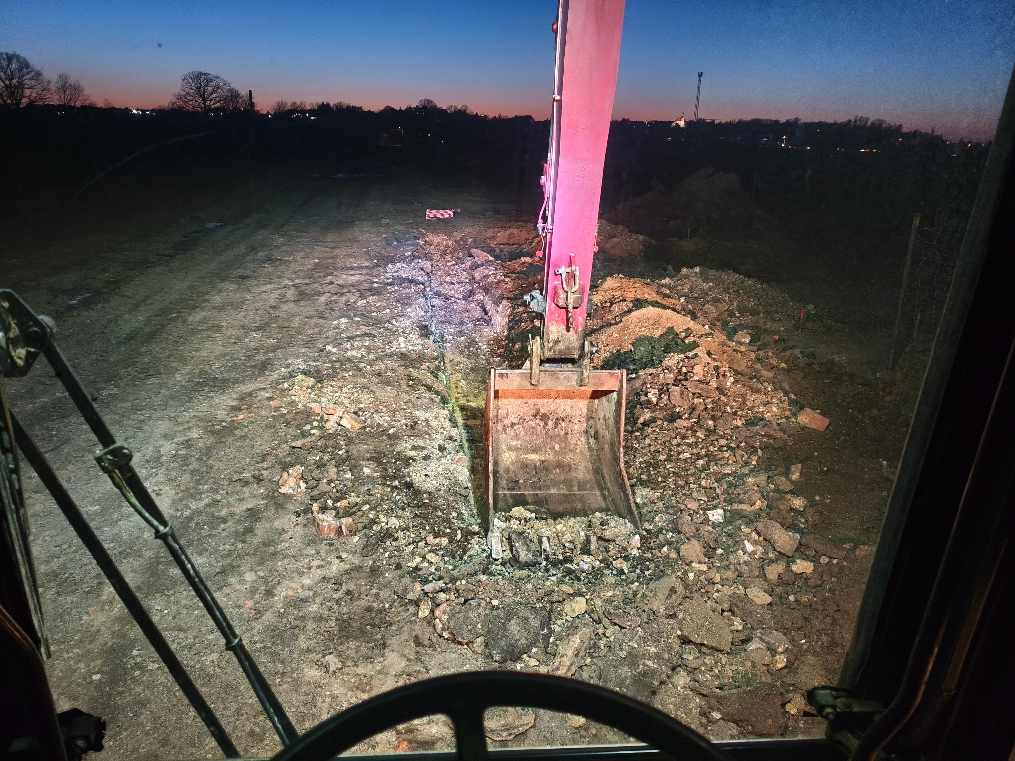 Widok z kabiny koparki na wykop w ziemi o zmierzchu. W tle widoczne zabudowania i drzewa. Na pierwszym planie łyżka koparki zagłębiająca się w gruz.