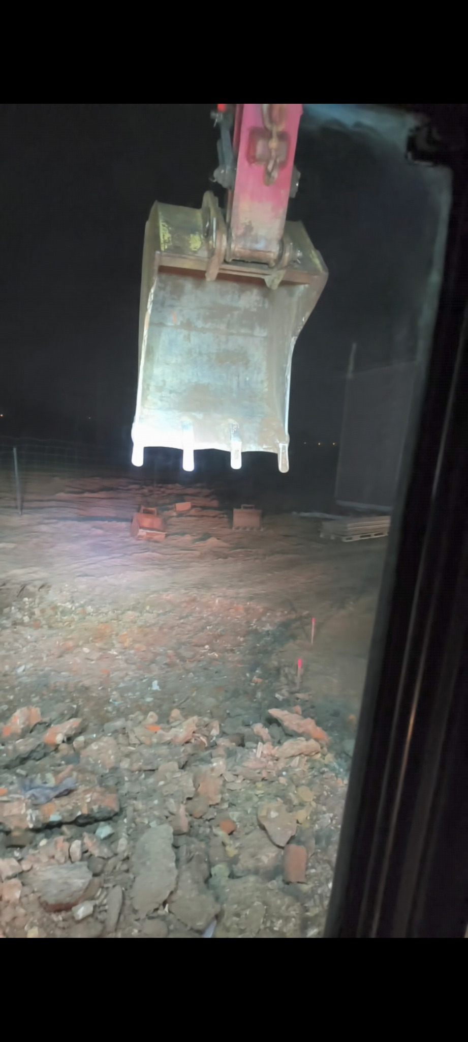 Łyżka koparki z zębami, widok z kabiny na gruzowisko w nocnym oświetleniu. Widoczne palety w tle i czerwone znaczniki w ziemi.