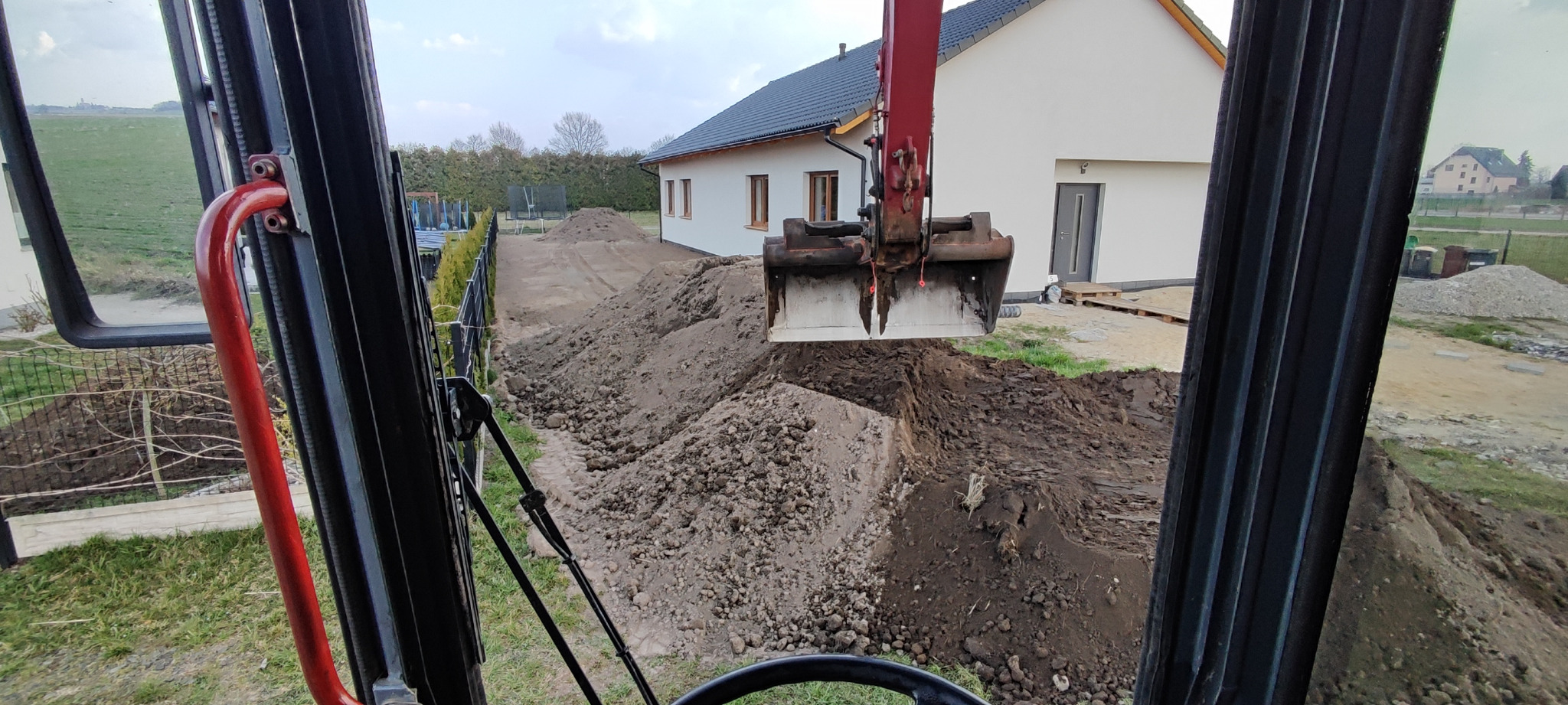 Widok z kabiny koparki na wykop wzdłuż ogrodzenia i domu. Łyżka koparki w trakcie pracy. Ślady opon na ziemi. Praca w toku, teren częściowo wyrównany.