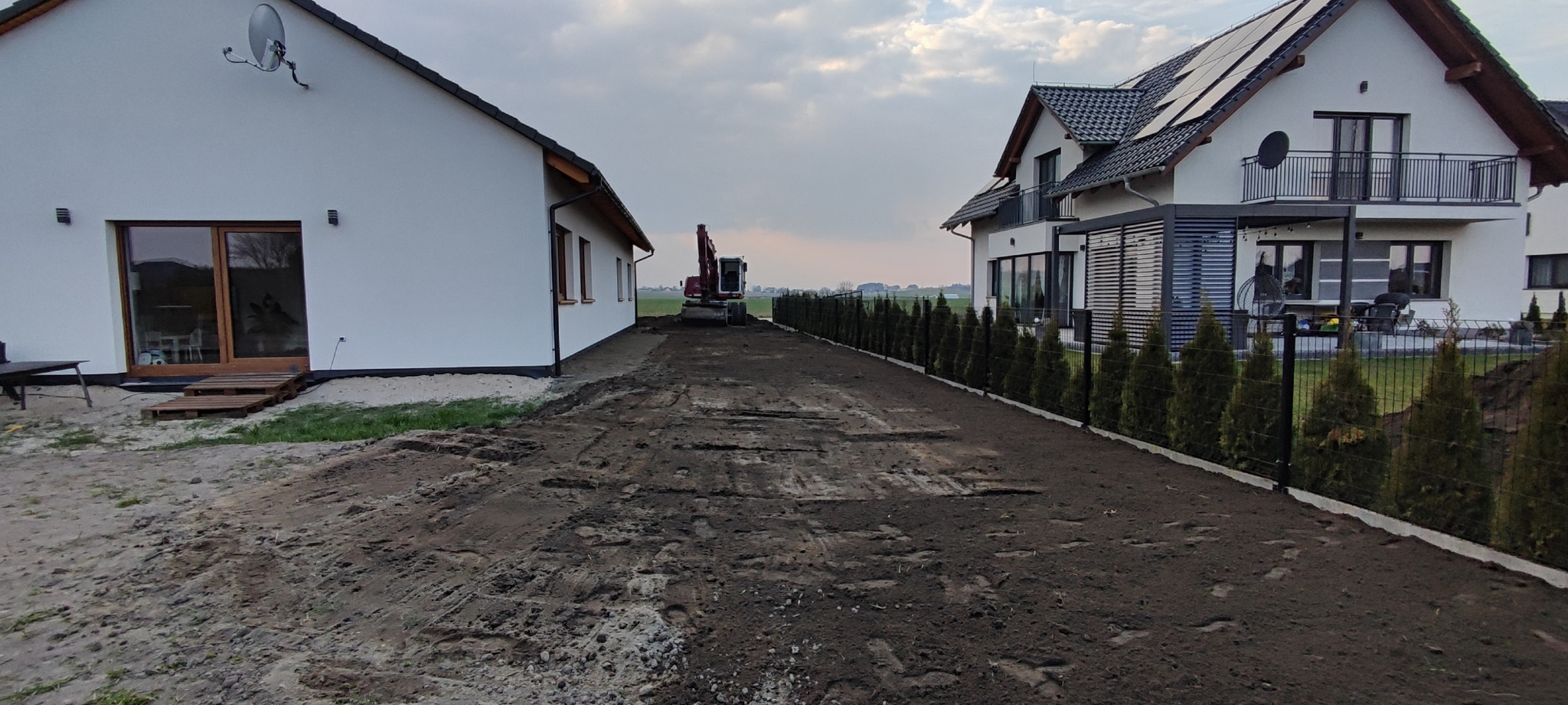 Niwelacja terenu pod budowę domu jednorodzinnego. W tle widoczny ciężki sprzęt budowlany. Uporządkowany teren z nasadzeniami po prawej stronie.