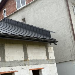 Dachy Kurek - Nowy, czarny dach z blachy na budynku w trakcie budowy, widoczne białe bloczki i szare spoiny. Rynna w kolorze dachu. Budynek sąsiaduje z innym, otynkowanym budynkiem.