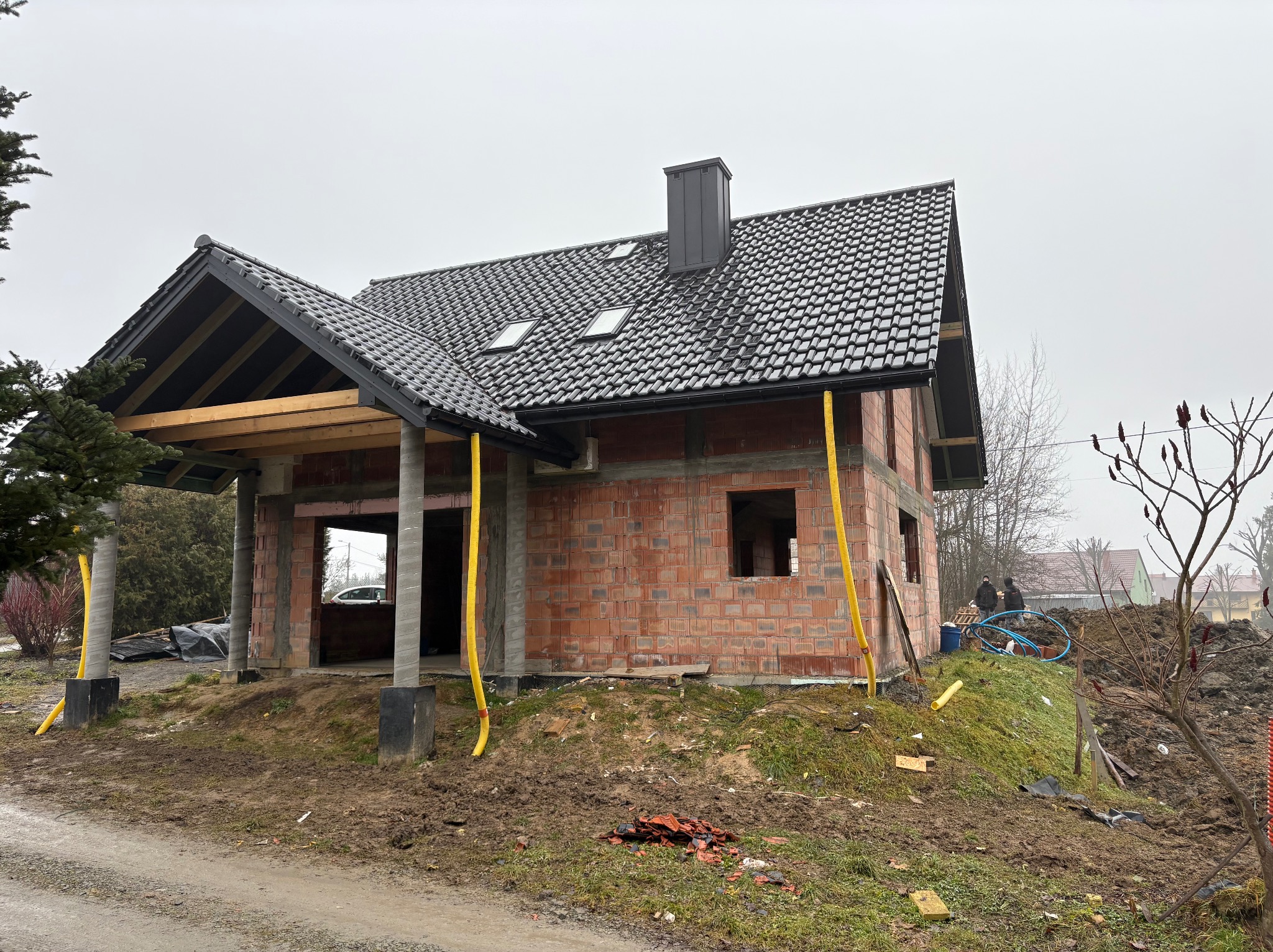 Surowy stan domu z ciemną dachówką karpiówką i oknami dachowymi. Widoczne elementy konstrukcyjne i fragmenty instalacji. Prace dekarskie w toku, stan surowy otwarty.