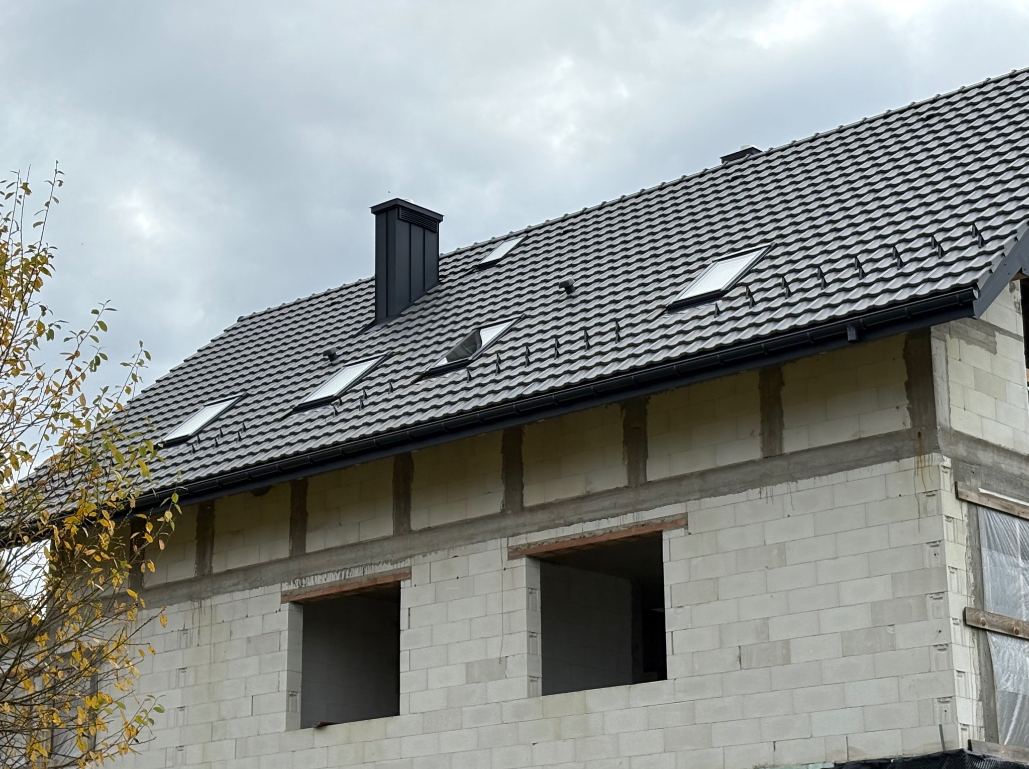 Dom w trakcie budowy z szarą dachówką, oknami dachowymi i czarnym kominem. Ściany z białych bloczków, widoczne otwory okienne. Częściowo widoczne drzewo z żółtymi liśćmi.