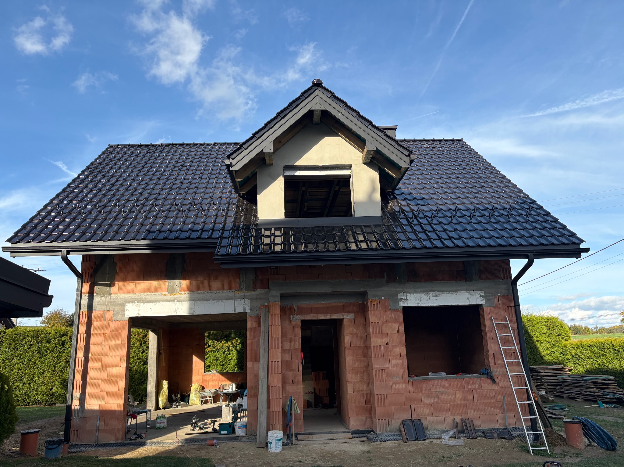 Dom w trakcie budowy z czarnym dachem z dachówki ceramicznej i oknem dachowym. Widoczne cegły i drabina. Błękitne niebo w tle.