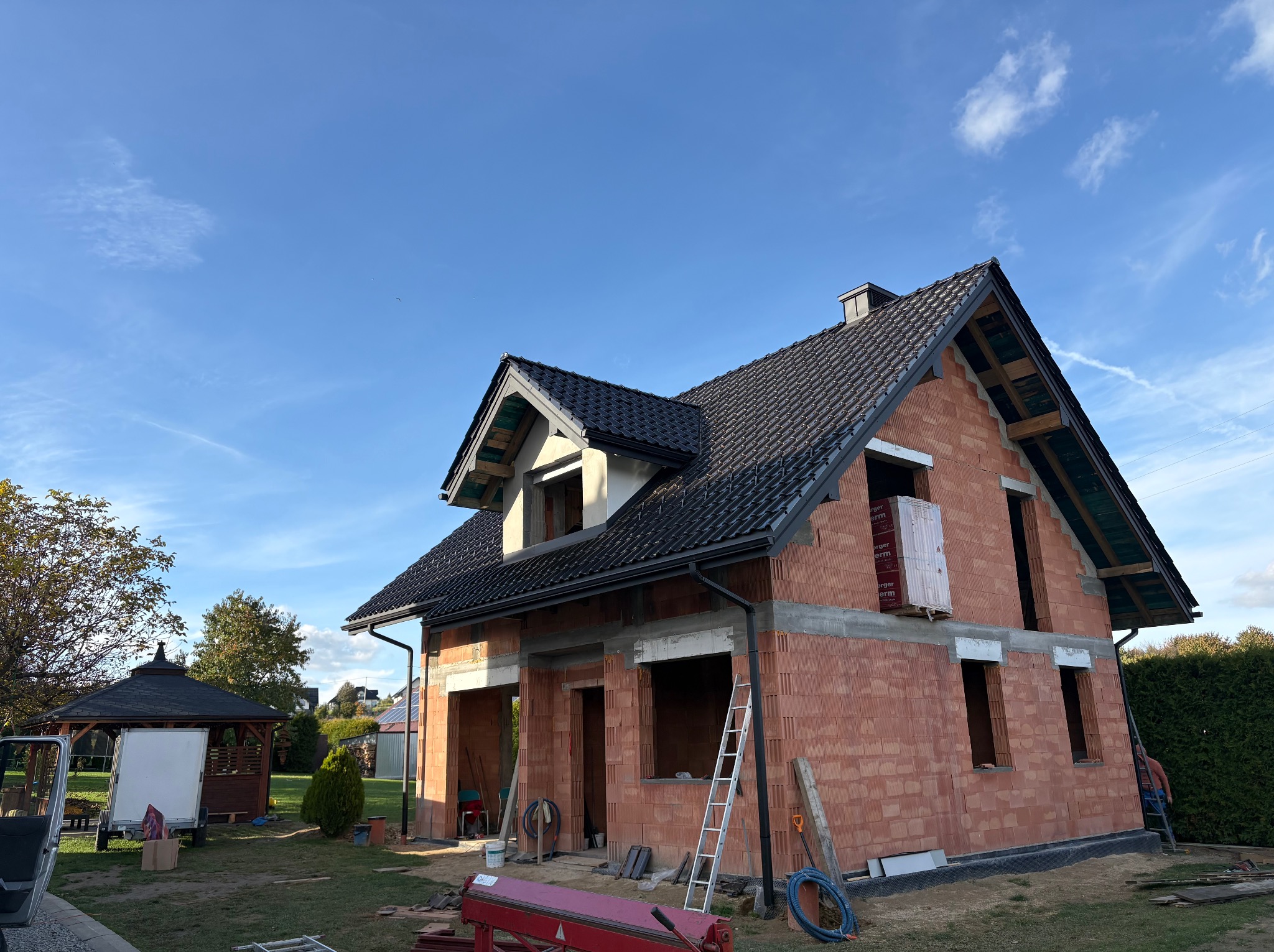 Dom w trakcie budowy z czarnym dachem z dachówki ceramicznej i zamontowanymi rynnami. Widoczne narzędzia i materiały budowlane. Błękitne niebo w tle.