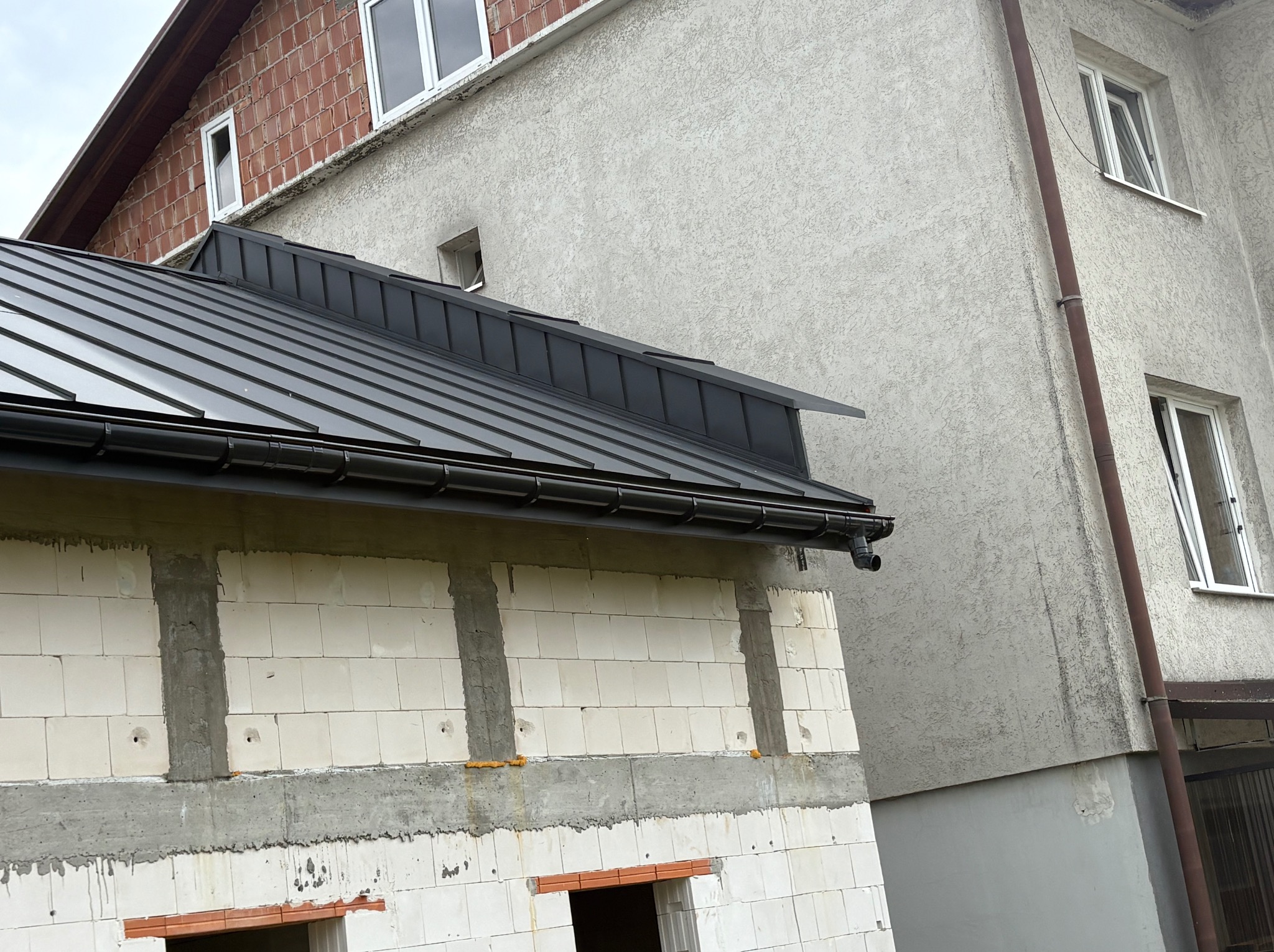 Nowy, czarny dach z blachy na budynku w trakcie budowy, widoczne białe bloczki i szare spoiny. Rynna w kolorze dachu. Budynek sąsiaduje z innym, otynkowanym budynkiem.