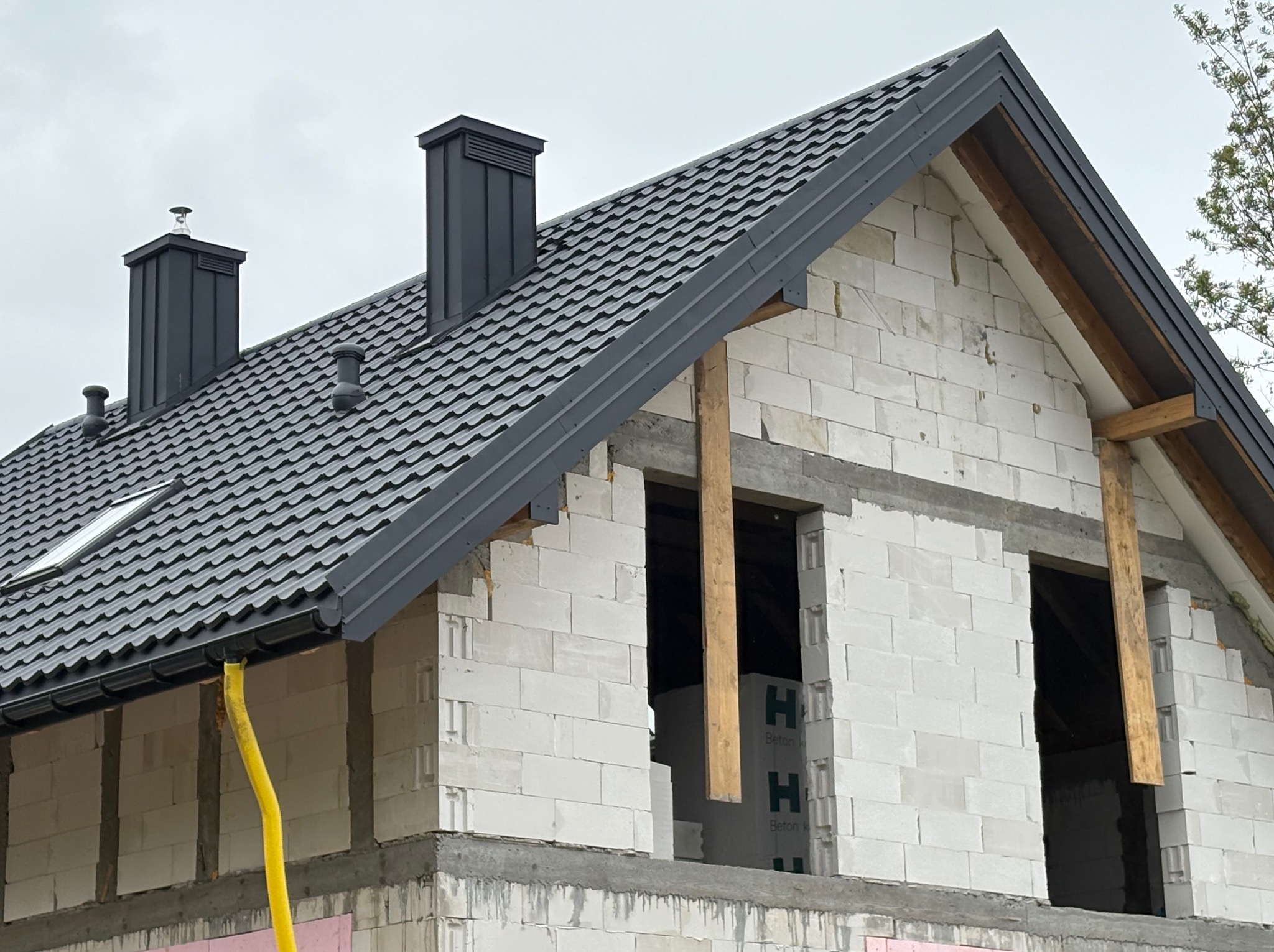 Dom w trakcie budowy z ciemnoszarą dachówką, widoczne kominy i rynna z żółtym wężem odprowadzającym wodę. Ściany z białego betonu komórkowego.