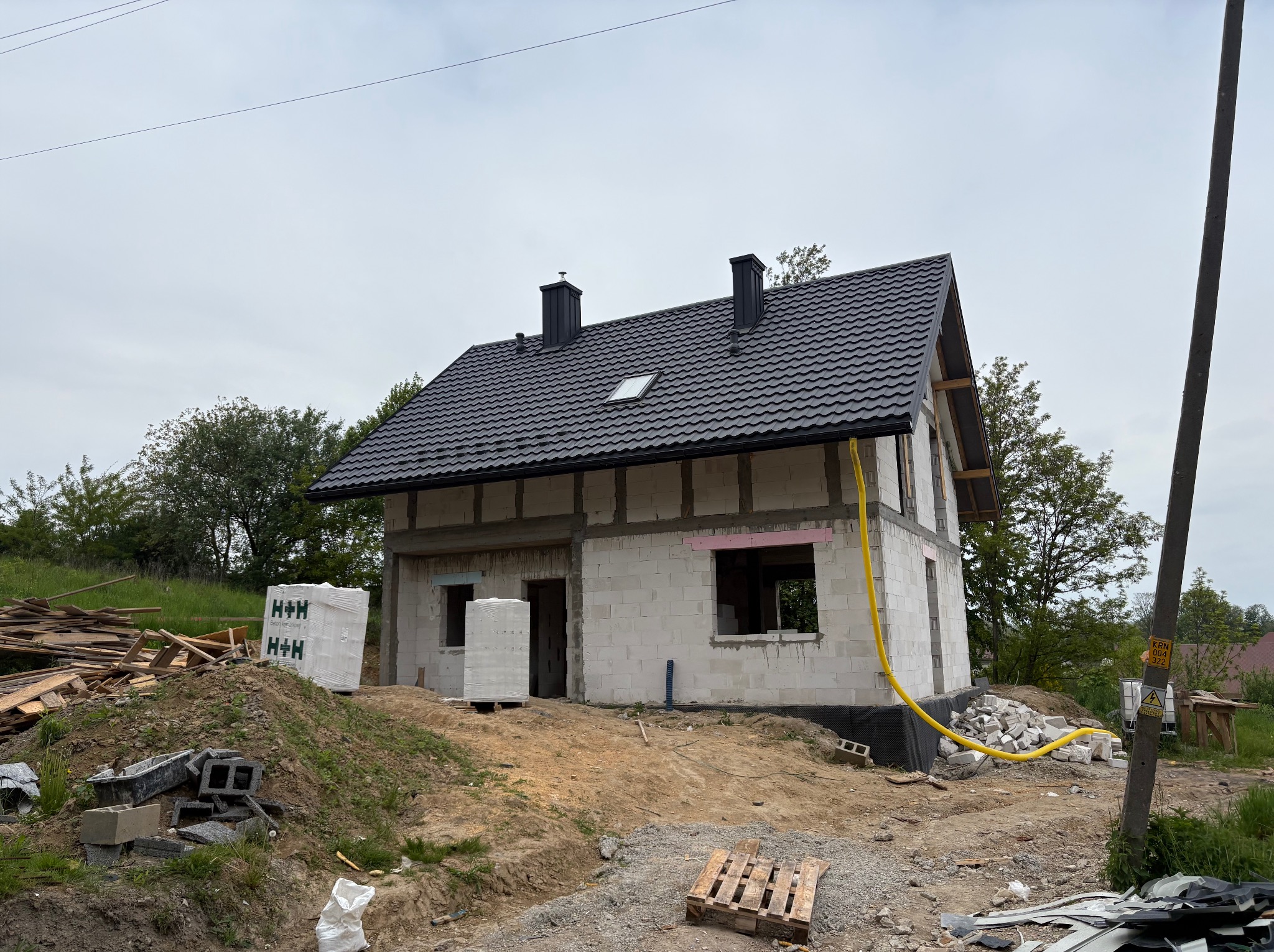 Dom w trakcie budowy z czarnym dachem z dachówki, widoczne kominy i okno dachowe. Na pierwszym planie materiały budowlane i teren budowy.