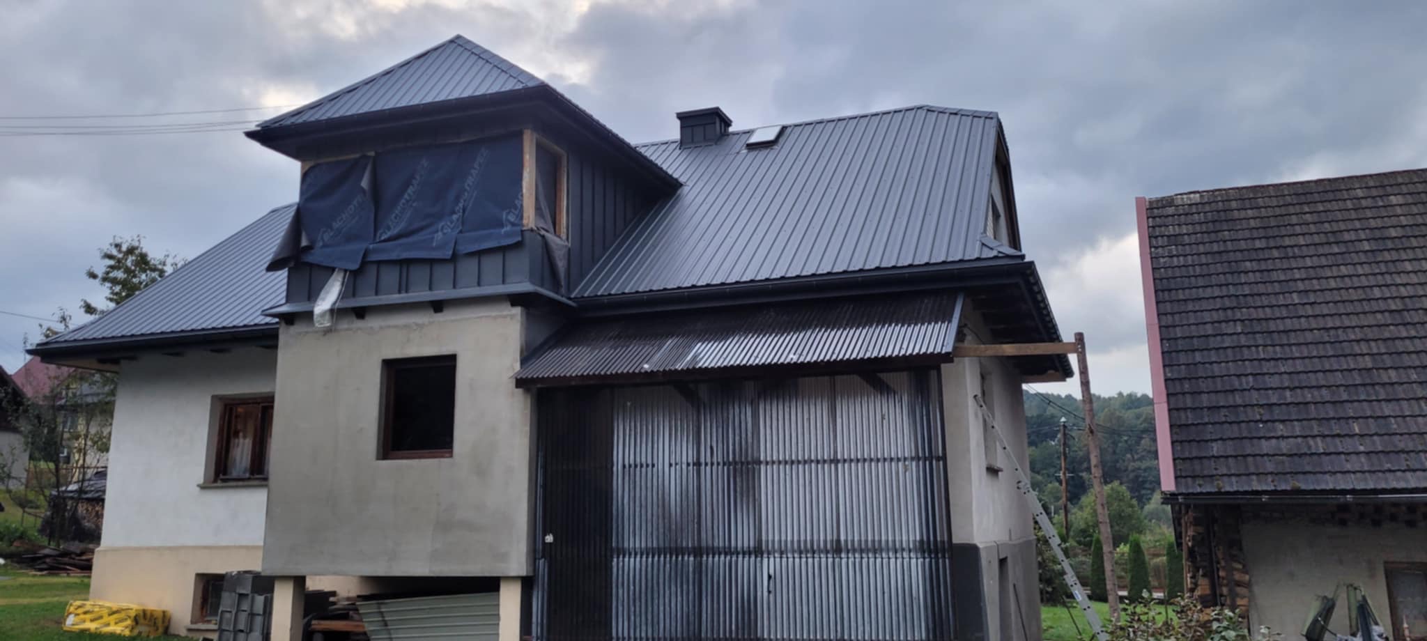 Dom w trakcie prac dekarskich: nowy, grafitowy dach z blachy trapezowej, okno dachowe zabezpieczone folią. Widoczna drabina i elementy konstrukcyjne.