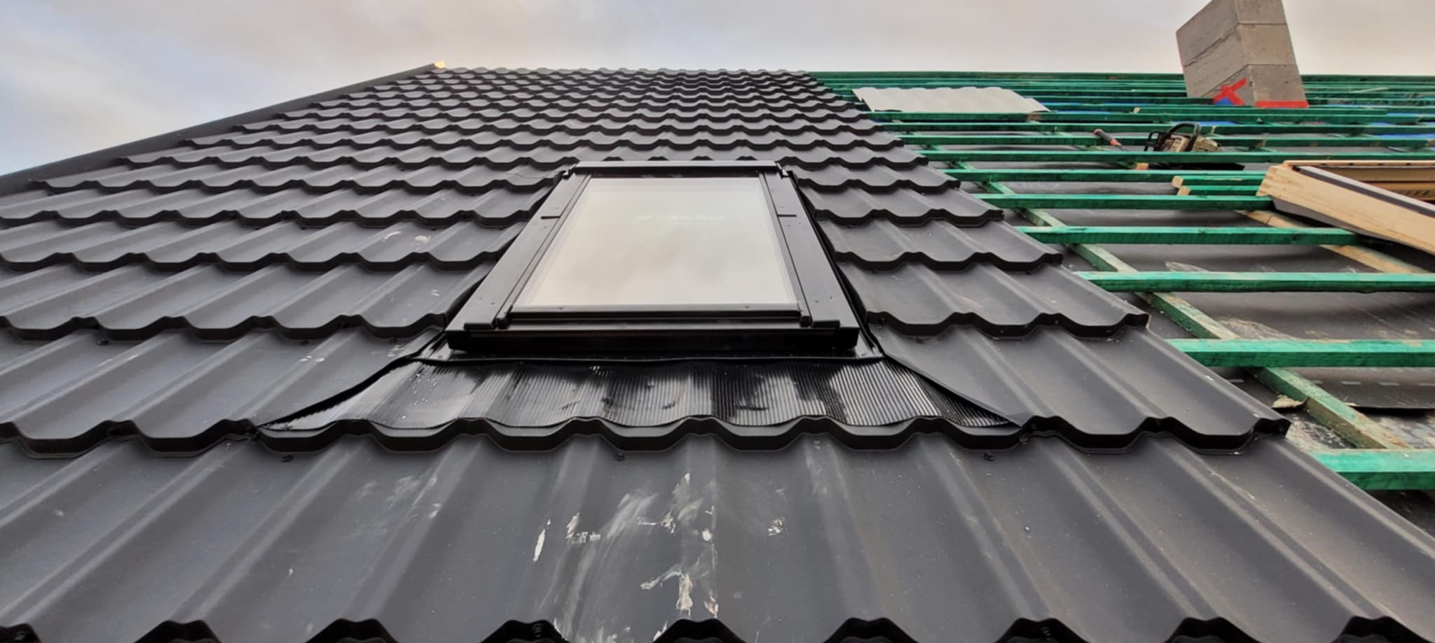 Częściowo pokryty dach z czarnej blachy trapezowej z oknem dachowym. Widoczna więźba dachowa i komin w trakcie budowy. Ujęcie z dołu pod kątem.