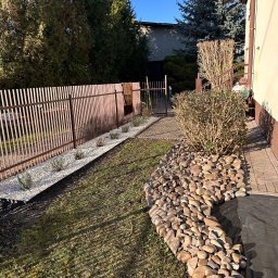 Sobolinhooo - Ogród po pielęgnacji: rabata z kamieniami, biały żwir wzdłuż brązowego ogrodzenia, świeżo posadzone rośliny, trawnik i krzewy. Słoneczny dzień.