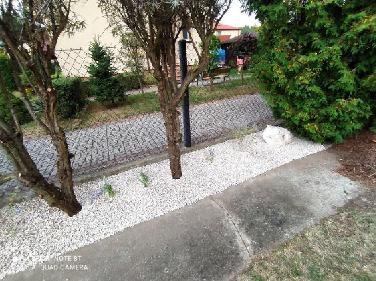 Obrzeże rabaty z białego kamienia oddzielone od trawnika betonową opaską, z widocznymi drzewami i tujami w tle, tworzące estetyczny element ogrodu.