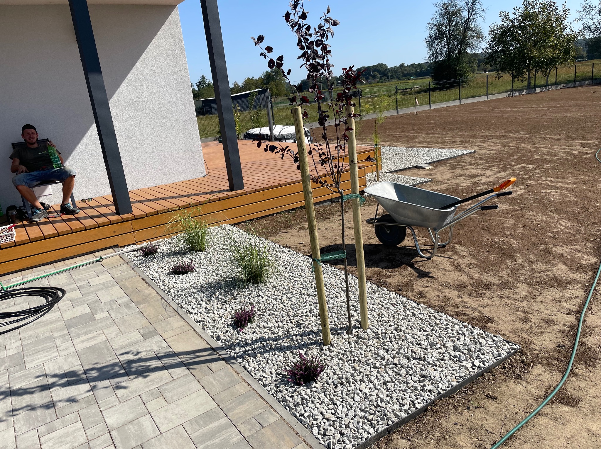 Nowoczesny ogród po realizacji: rabata z białym grysem, ozdobne trawy i drzewko. W tle trawnik, wózek ogrodowy i siedzący mężczyzna.