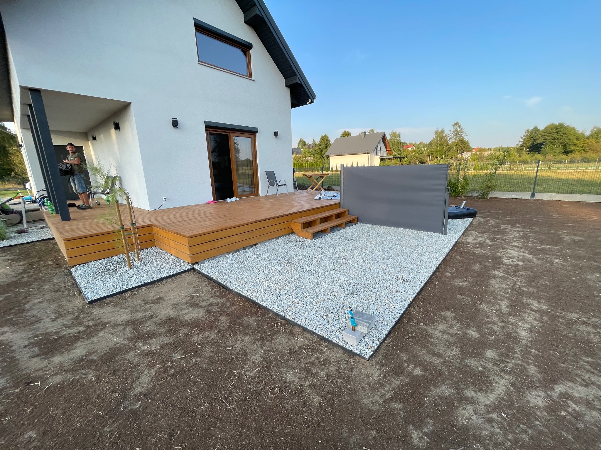 Nowoczesny taras z drewnianą podłogą i białym żwirem, widok z perspektywy ogrodu. Minimalistyczny design, jasne kolory, świeżo założony trawnik.