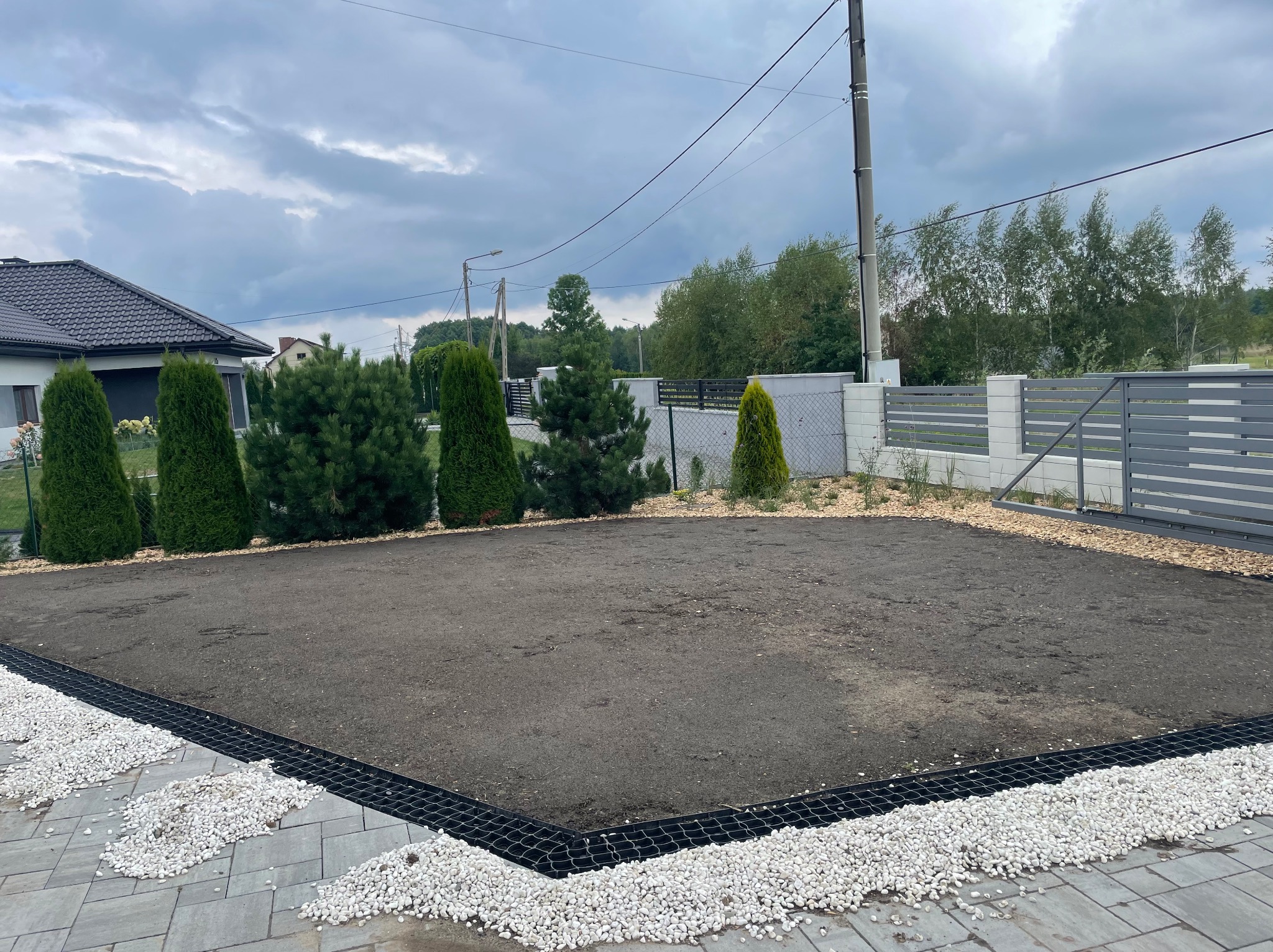 Przygotowany teren pod trawnik z rolki, obrzeża z białego kamienia i siatki. W tle iglaki i ogrodzenie posesji. Pochmurne niebo.