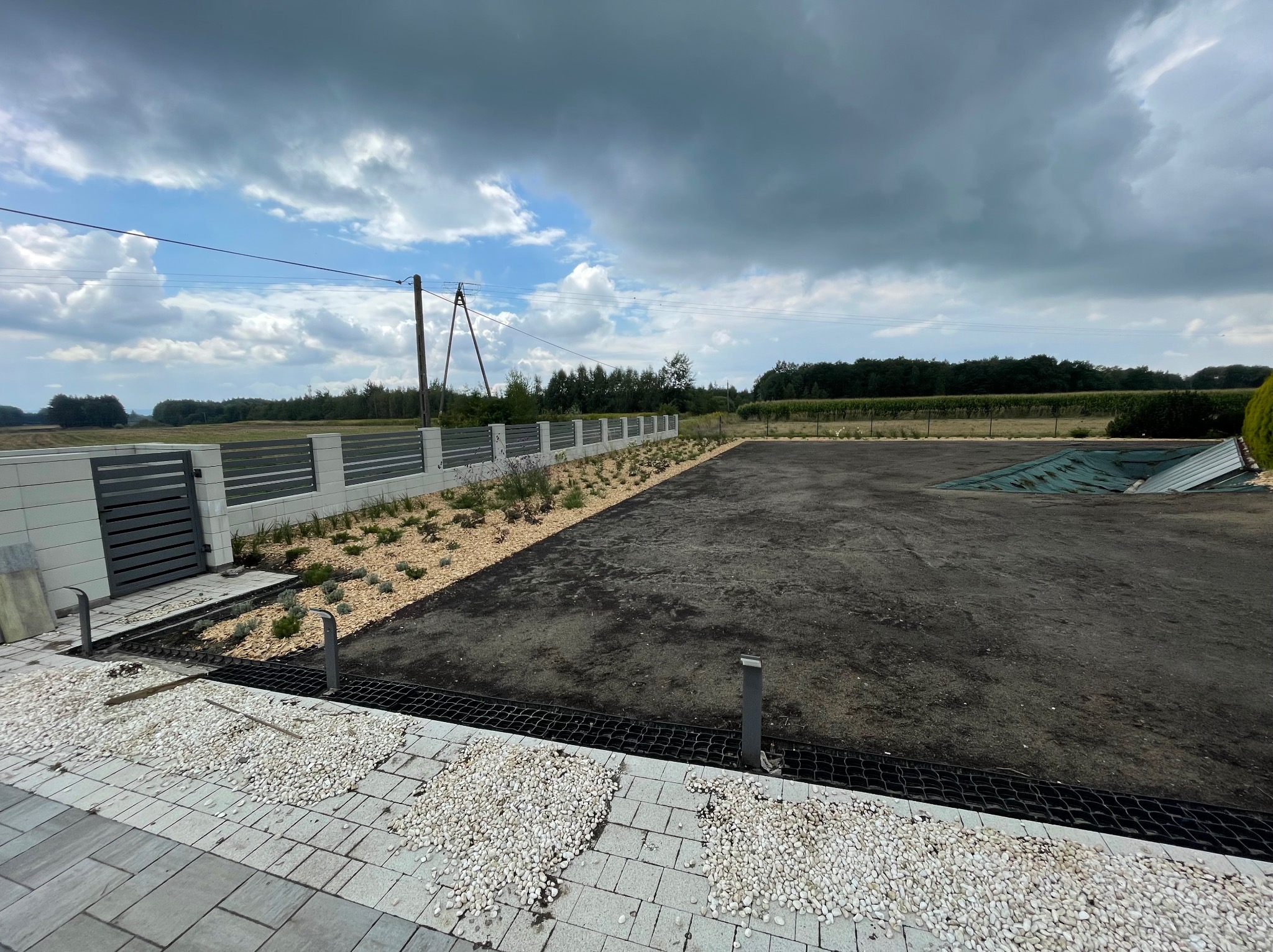 Przygotowany teren pod trawnik z rolki, obrzeża z białego kamienia i rabaty z korą, ogrodzenie panelowe w tle, pochmurne niebo.