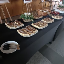 AGMAR SPÓŁKA Z OGRANICZONĄ ODPOWIEDZIALNOŚCIĄ - Stół cateringowy z sernikiem w papilotkach, deserami w szklankach i kwiatami. Widoczne szczypce do nakładania. Czarny obrus, jasne tło.