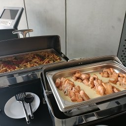 AGMAR SPÓŁKA Z OGRANICZONĄ ODPOWIEDZIALNOŚCIĄ - Bufet cateringowy z penne w sosie pomidorowym i kawałkami łososia w kremowym sosie, gotowy do serwowania. Na pierwszym planie biały talerz z łyżką i łopatką.