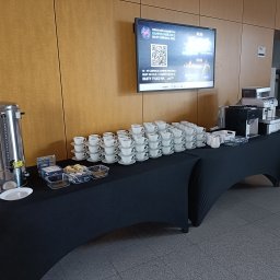 AGMAR SPÓŁKA Z OGRANICZONĄ ODPOWIEDZIALNOŚCIĄ - Stół cateringowy z piramidą filiżanek, automatami do kawy i przekąskami, przygotowany na wydarzenie firmowe z grillem UAM w tle na ekranie.