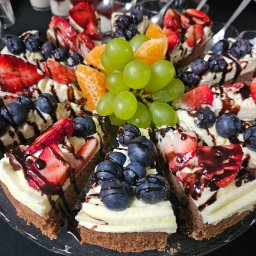 AGMAR SPÓŁKA Z OGRANICZONĄ ODPOWIEDZIALNOŚCIĄ - Apetyczny tort owocowy na catering: truskawki, borówki, mandarynki i winogrona, polany czekoladą, na przezroczystym talerzu. Idealny na firmowe przyjęcie.