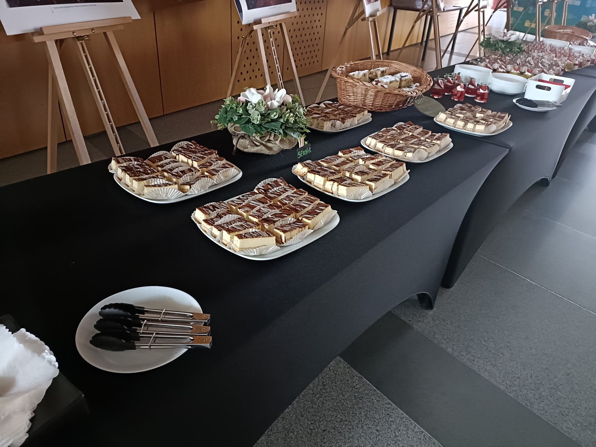 Stół cateringowy z sernikiem w papilotkach, deserami w szklankach i kwiatami. Widoczne szczypce do nakładania. Czarny obrus, jasne tło.