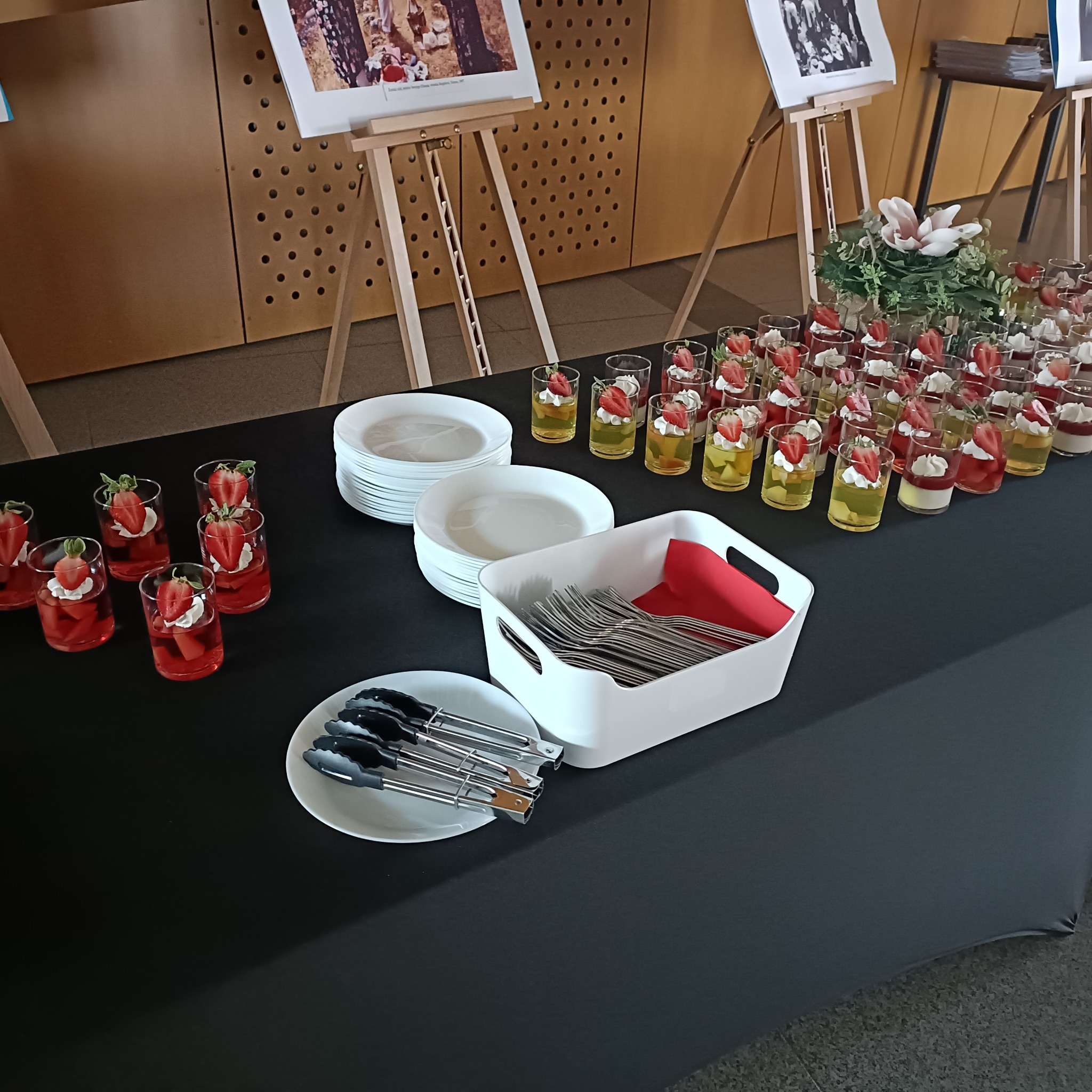 Stół cateringowy z deserami w szklankach, sztućcami i kwiatami. Widoczne zdjęcia na sztalugach w tle. Przygotowanie do imprezy firmowej lub świątecznej.