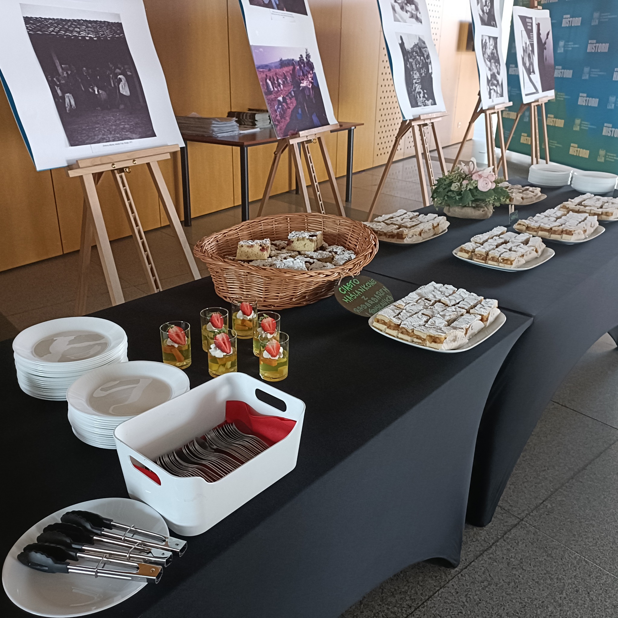 Stół cateringowy z ciastami, deserami i napojami, ustawiony przed ekspozycją fotografii na sztalugach. Widoczne talerze, sztućce i dekoracje kwiatowe.