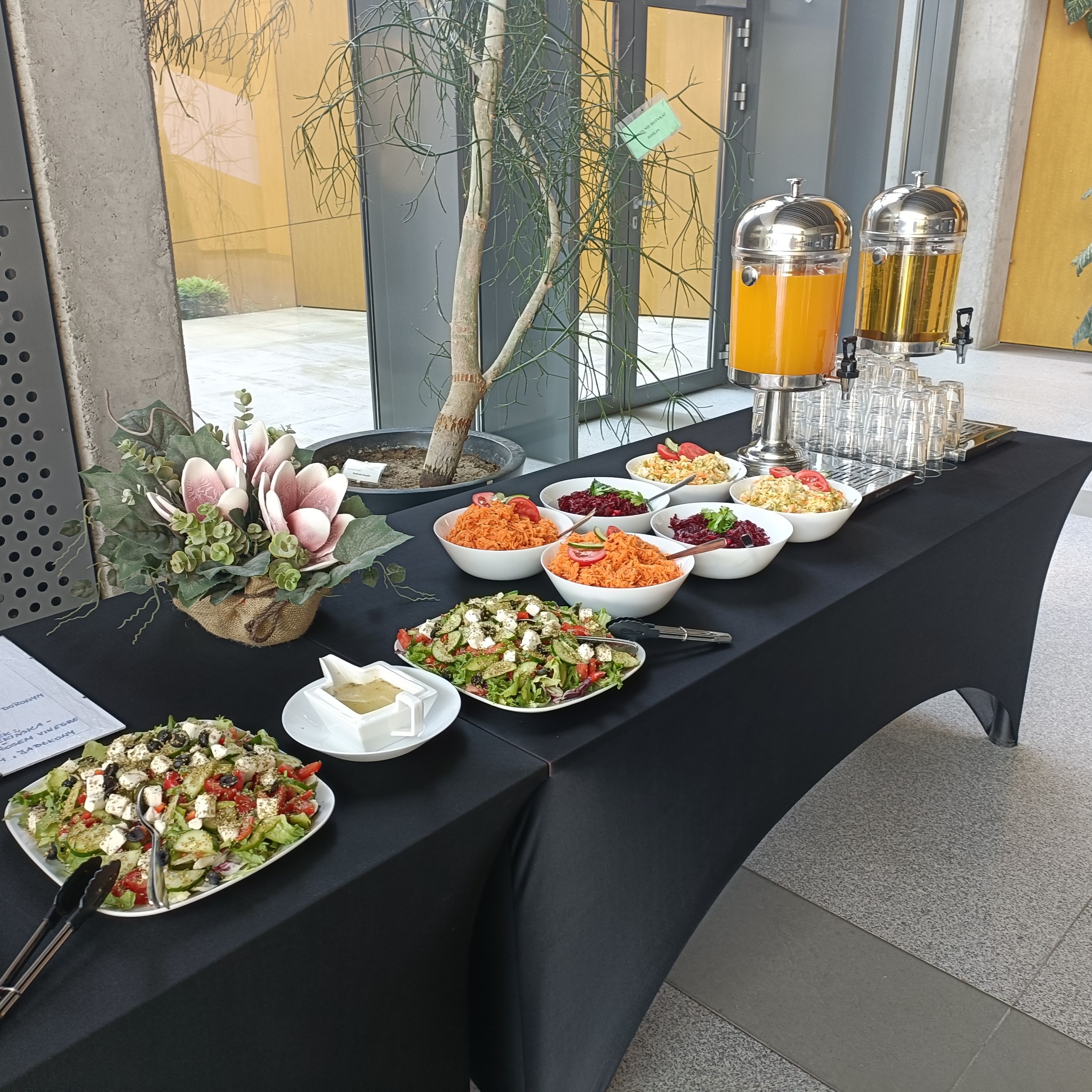Stół cateringowy z sałatkami, surówkami i napojami w dyspenserach. Dekoracja kwiatowa i roślina doniczkowa w tle. Czarny obrus.