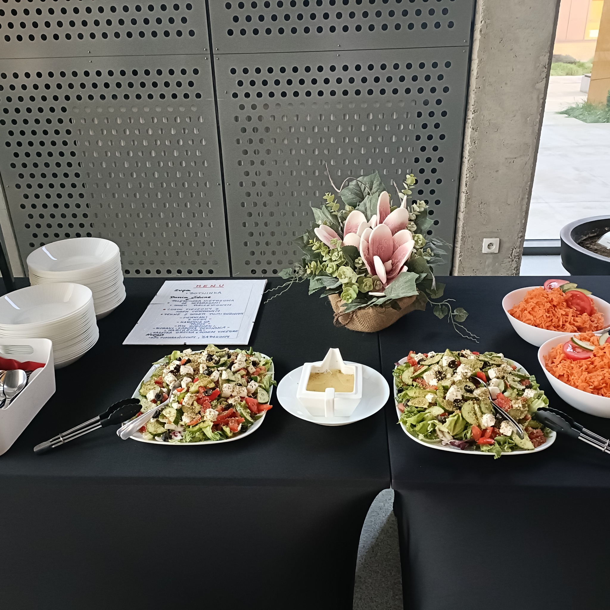 Stół cateringowy z sałatkami greckimi, surówką z marchwi, stosem talerzy i menu na czarnym obrusie, dekoracja kwiatowa w tle.