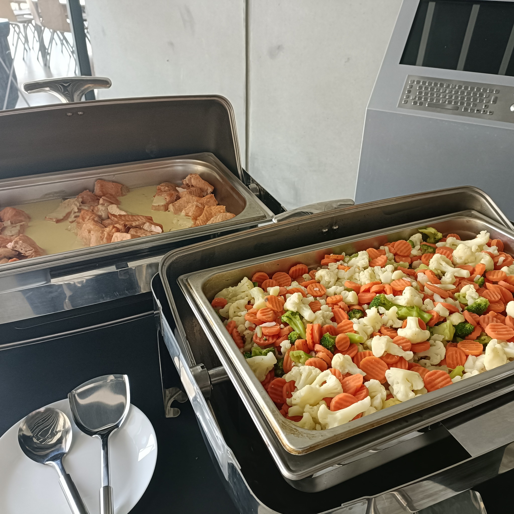 Dwa metalowe bemary z gotowanymi warzywami (brokuły, marchew, kalafior) i łososiem w sosie, ustawione na czarnym stole, obok biały talerz i łyżki do nakładania.