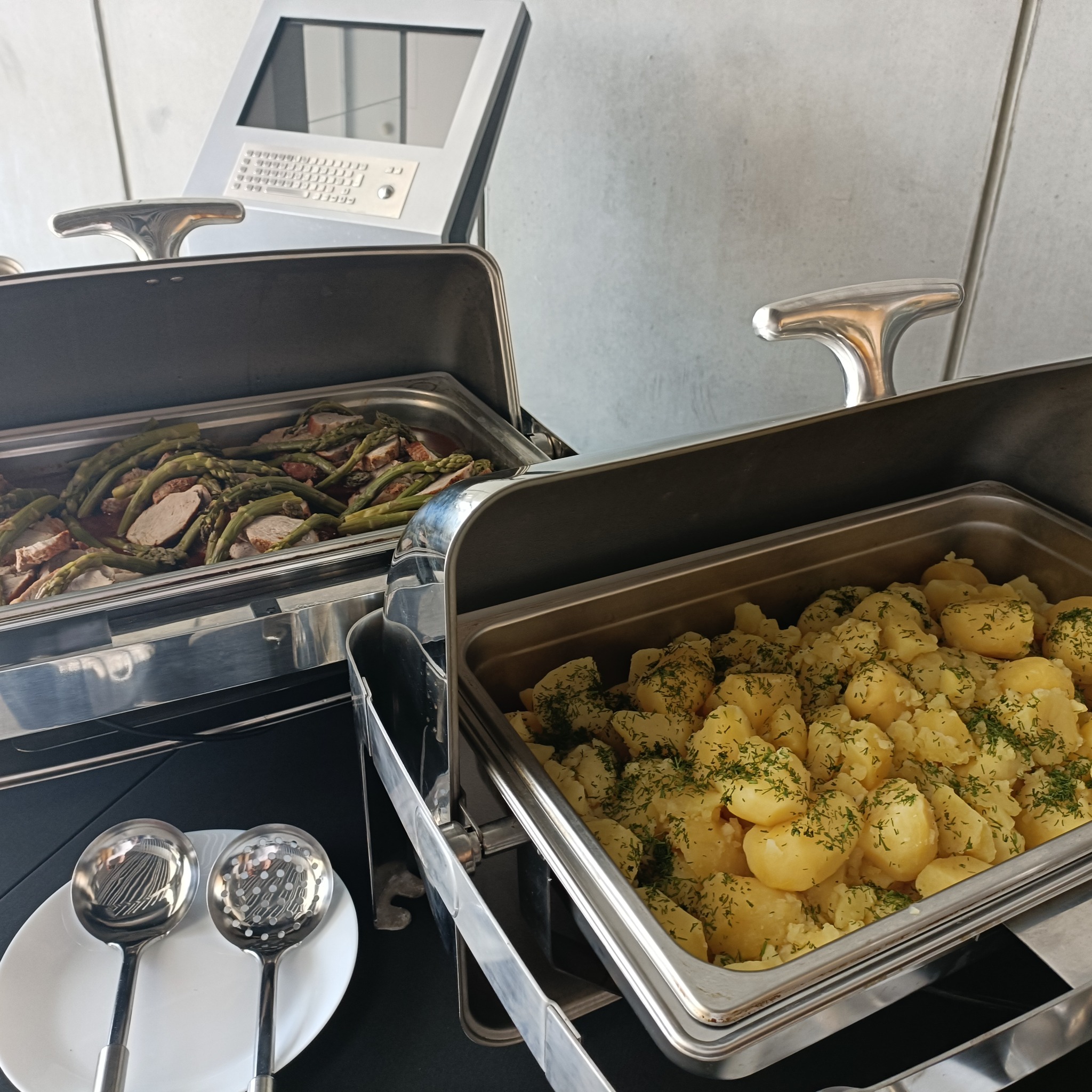 Dwa bemary ze srebrnymi pokrywami: w jednym mięso z zielonymi szparagami, w drugim ziemniaki z koperkiem. Obok biały talerz z łyżkami cedzakowymi.