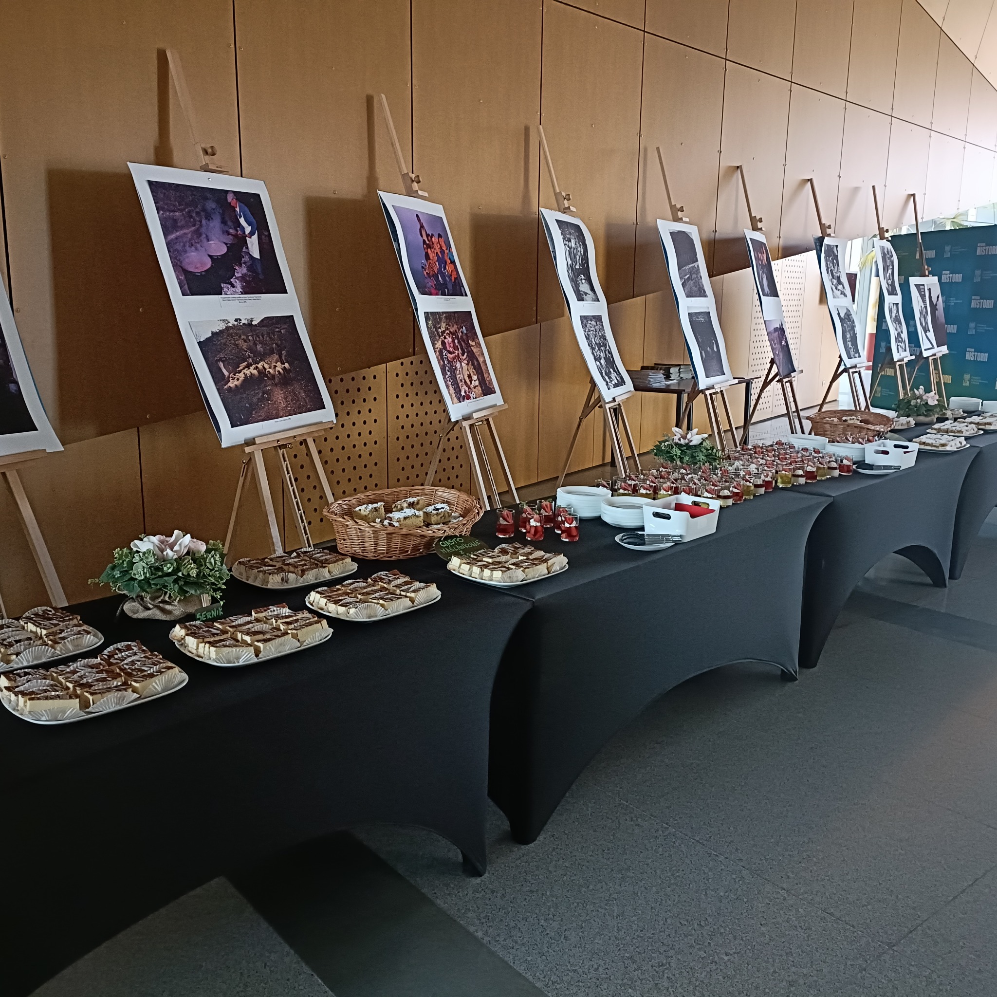 Stół cateringowy z deserami i zdjęciami na sztalugach w tle. Widoczne ciasta, desery w szklankach i koszyk z wypiekami. Czarny obrus, jasne tło.