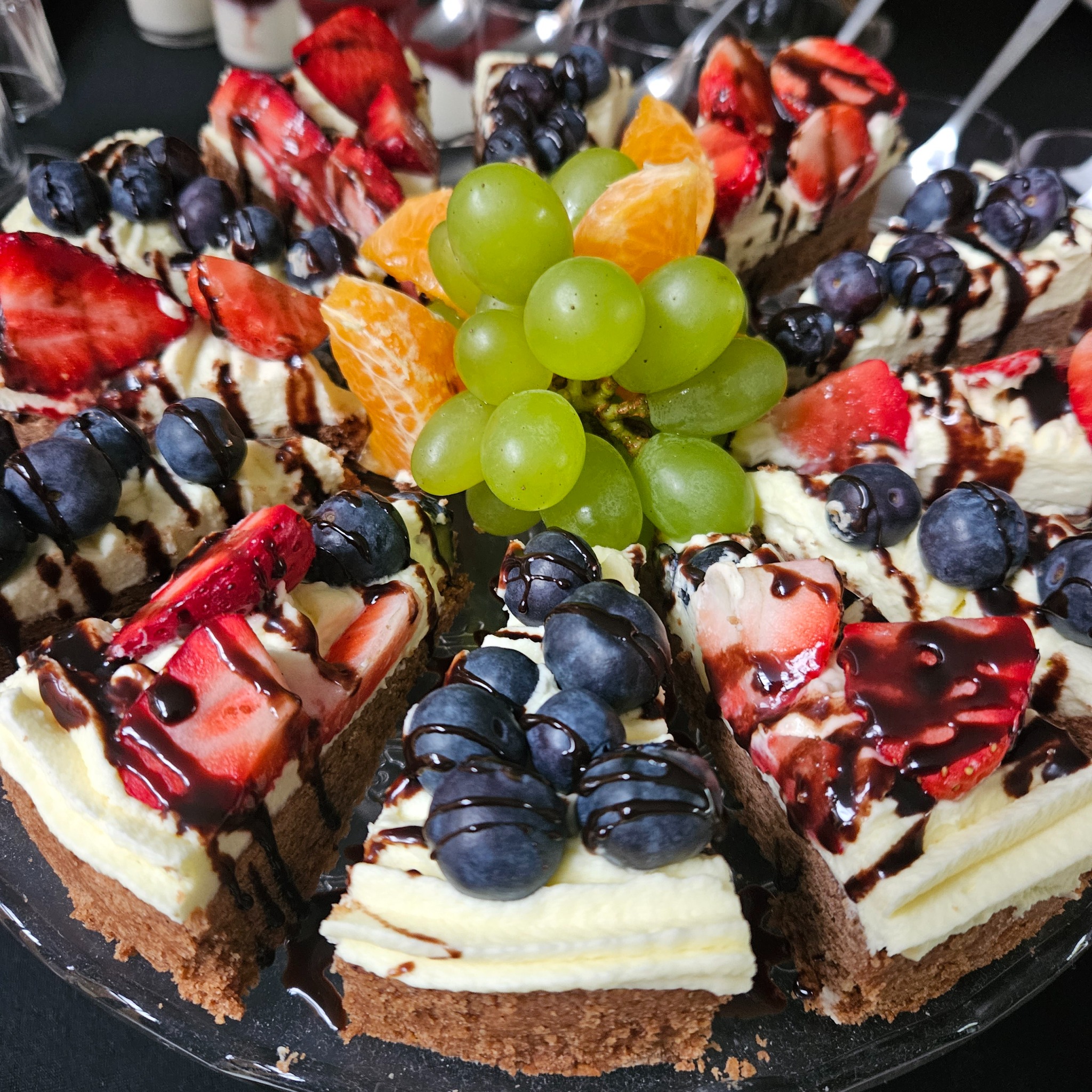 Apetyczny tort owocowy na catering: truskawki, borówki, mandarynki i winogrona, polany czekoladą, na przezroczystym talerzu. Idealny na firmowe przyjęcie.