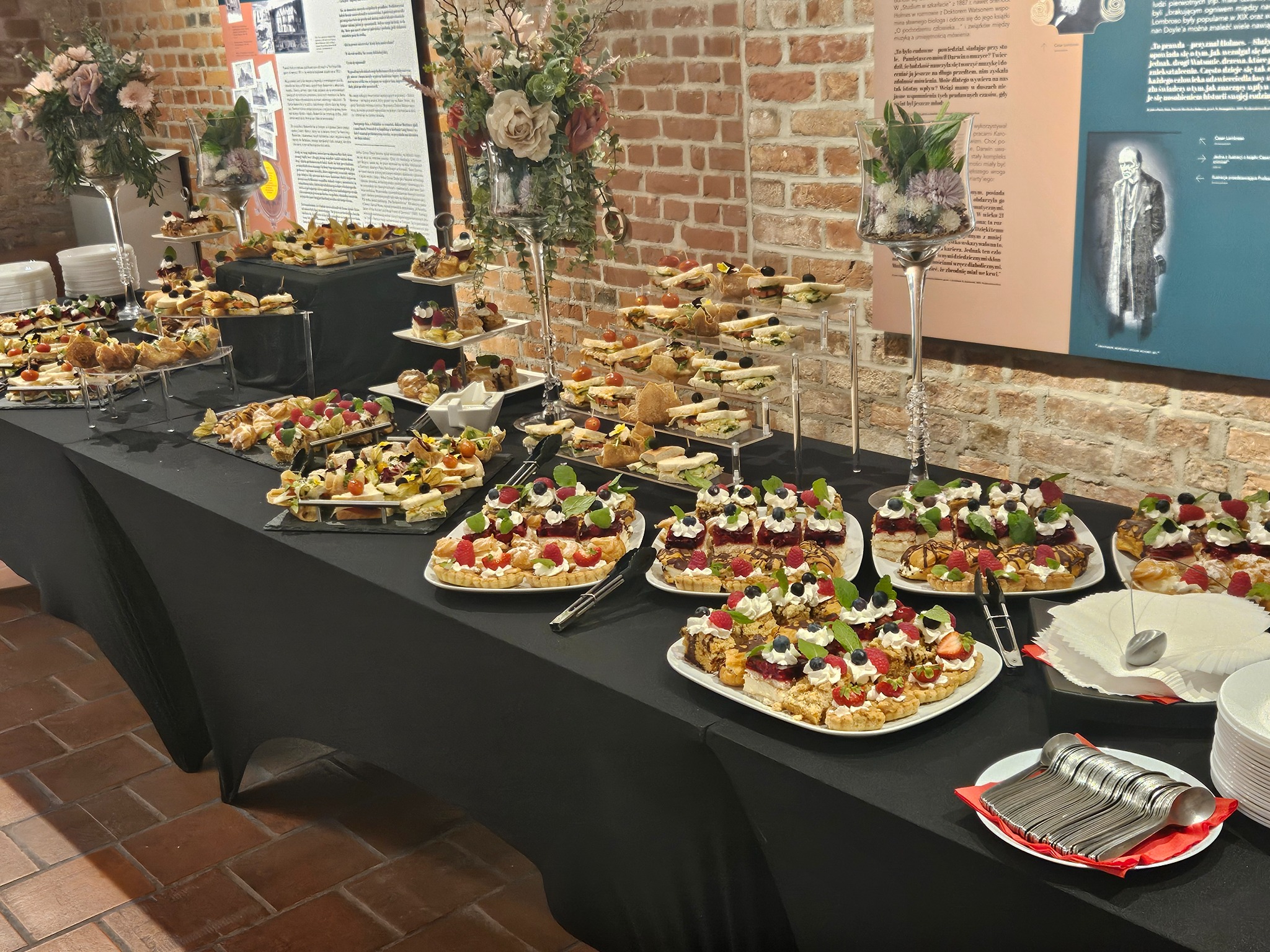 Stół cateringowy z różnorodnymi przekąskami: tartaletki z owocami, kanapki i desery. Dekoracje kwiatowe i elegancka zastawa. Tło z ceglanej ściany i ekspozycji.