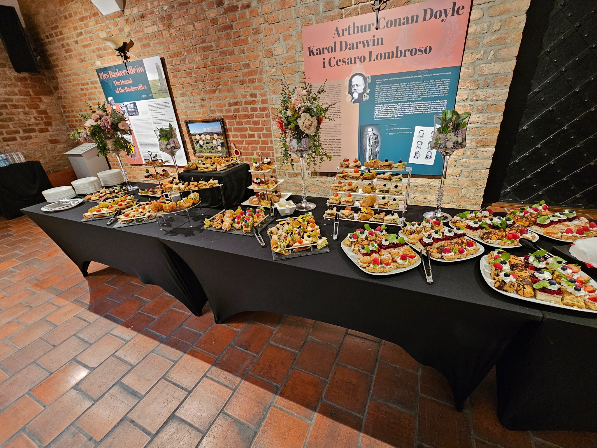 Stół cateringowy z przekąskami, dekoracjami kwiatowymi i plakatami w tle, ustawiony w ceglanym wnętrzu. Różnorodność kanapek, tartaletek i ciast.
