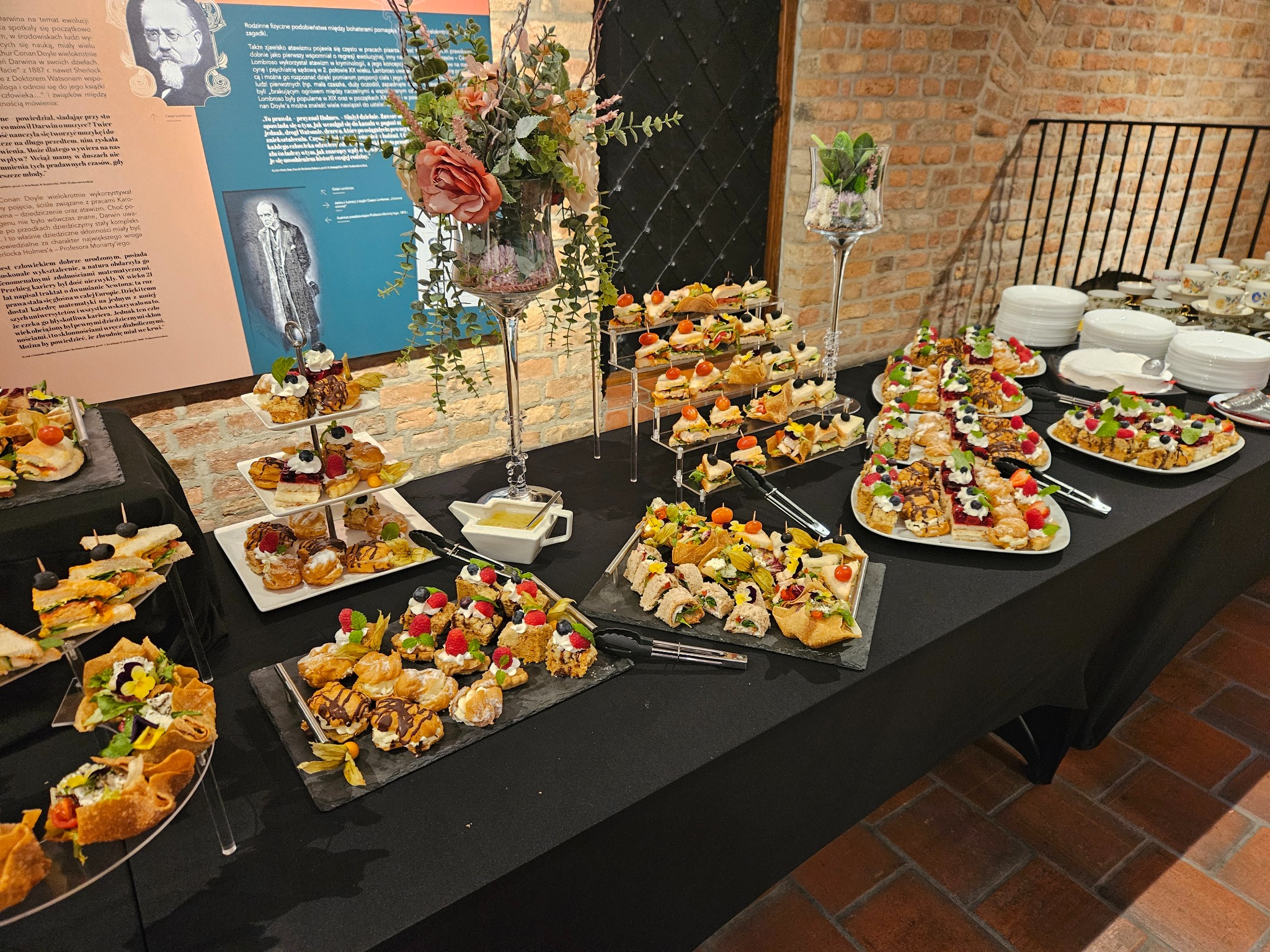 Bogato zastawiony stół cateringowy z różnorodnymi przekąskami, ciastami i dekoracjami kwiatowymi, idealny na eleganckie przyjęcie firmowe lub świąteczne.