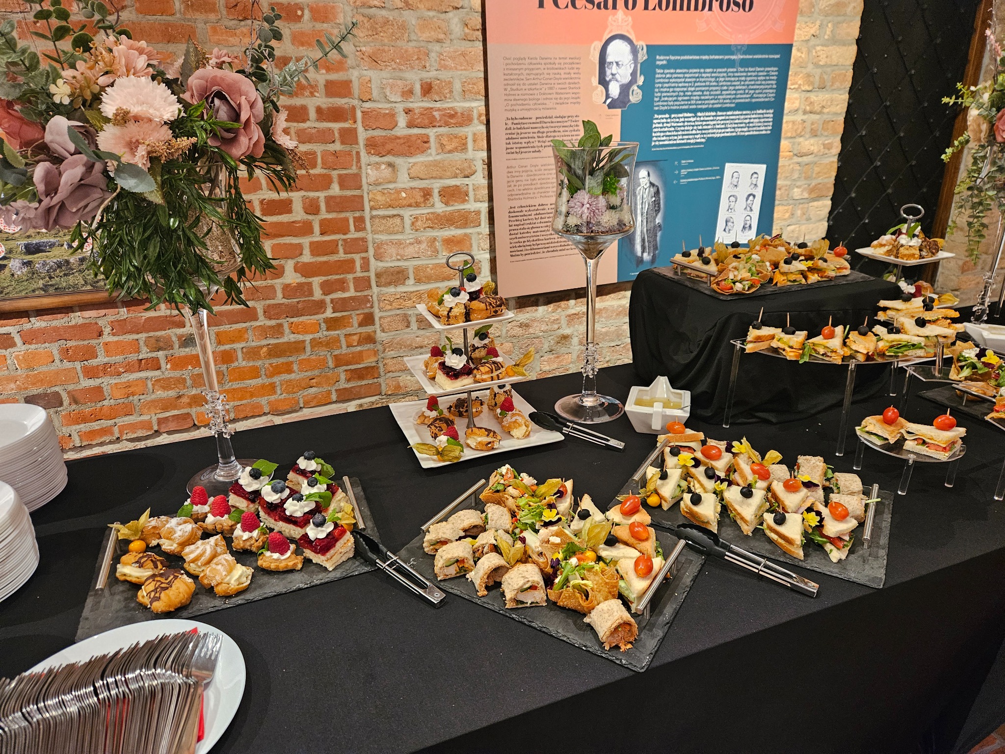 Stół cateringowy z różnorodnymi przekąskami: mini kanapeczki, roladki, ciastka, owoce. Dekoracje kwiatowe i elegancka zastawa. Ceglana ściana w tle.