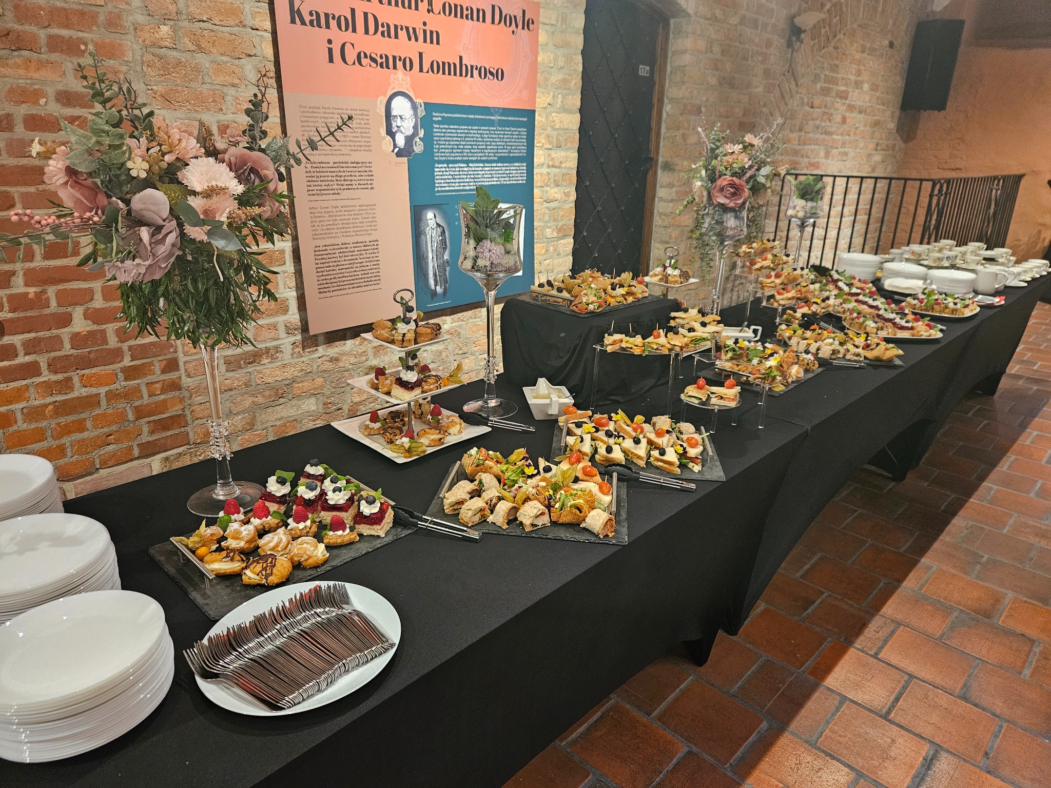 Stół cateringowy z przekąskami, dekoracjami kwiatowymi i stertami talerzy w ceglanym wnętrzu. Różnorodne kanapki, desery i napoje na czarnym obrusie.