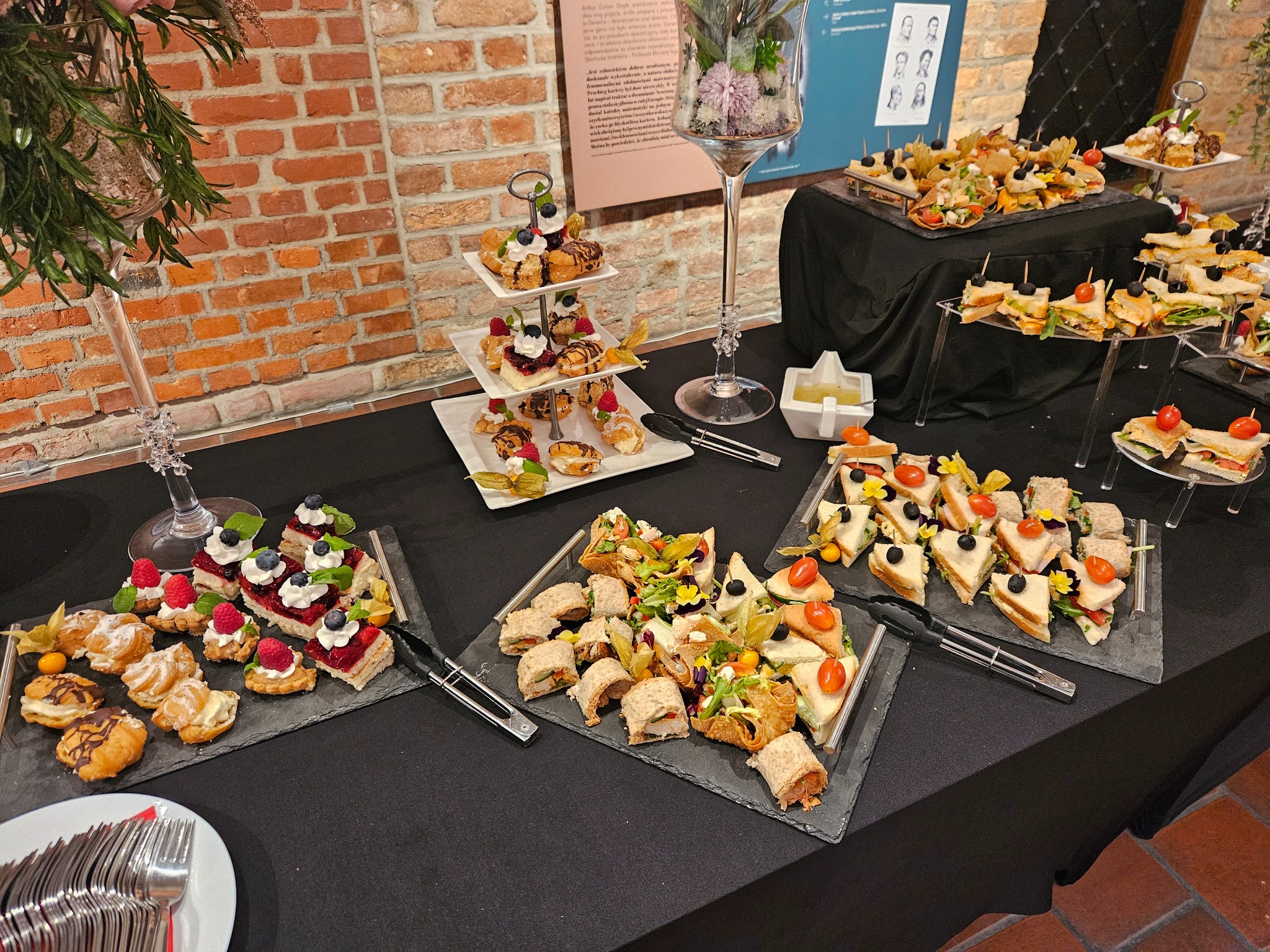 Elegancki stół cateringowy z różnorodnymi przekąskami: mini torty, kanapki i roladki, idealne na firmowe przyjęcie lub świąteczne spotkanie. Dekoracje kwiatowe i ceglana ściana w tle.