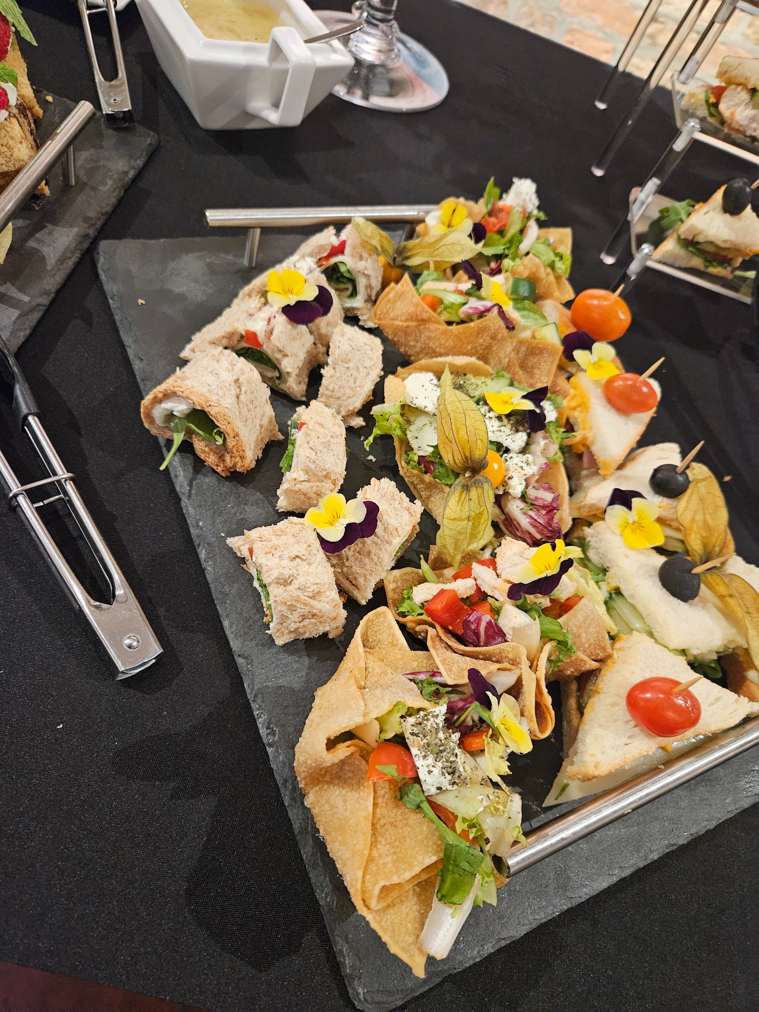 Elegancki catering: kanapki, roladki i sałatki w kruchych koszyczkach, udekorowane jadalnymi kwiatami, podane na czarnym, łupkowym talerzu.