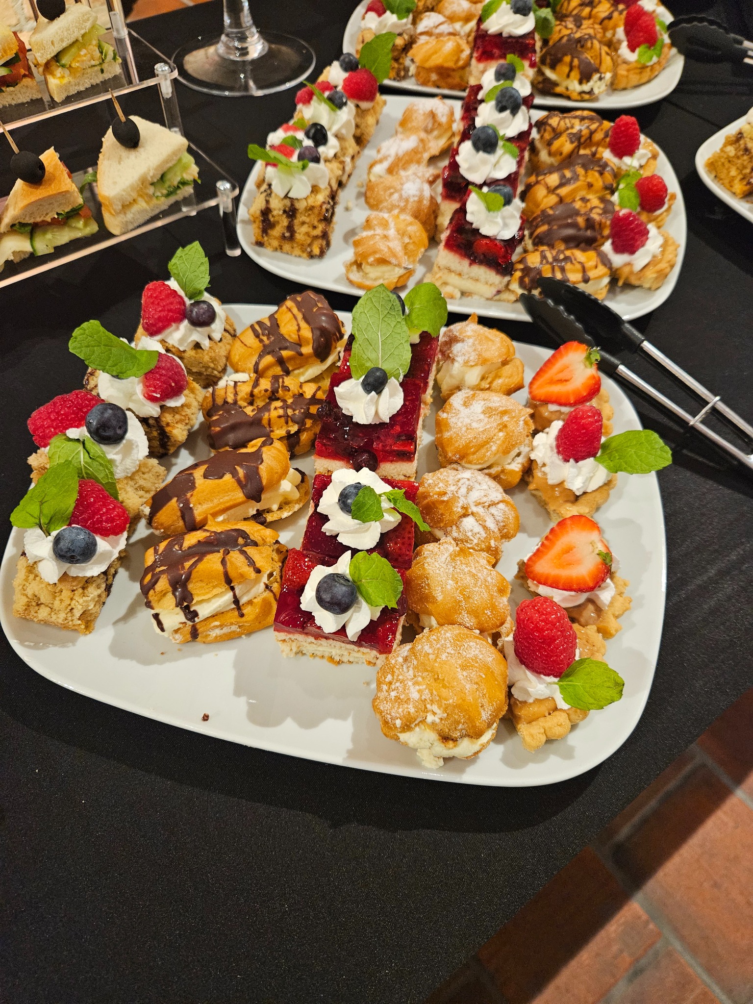 Elegancki catering: taca z mini deserami (eklerki, ciasta) udekorowanymi owocami (maliny, borówki) i miętą, obok szczypce i kanapki na stojakach.