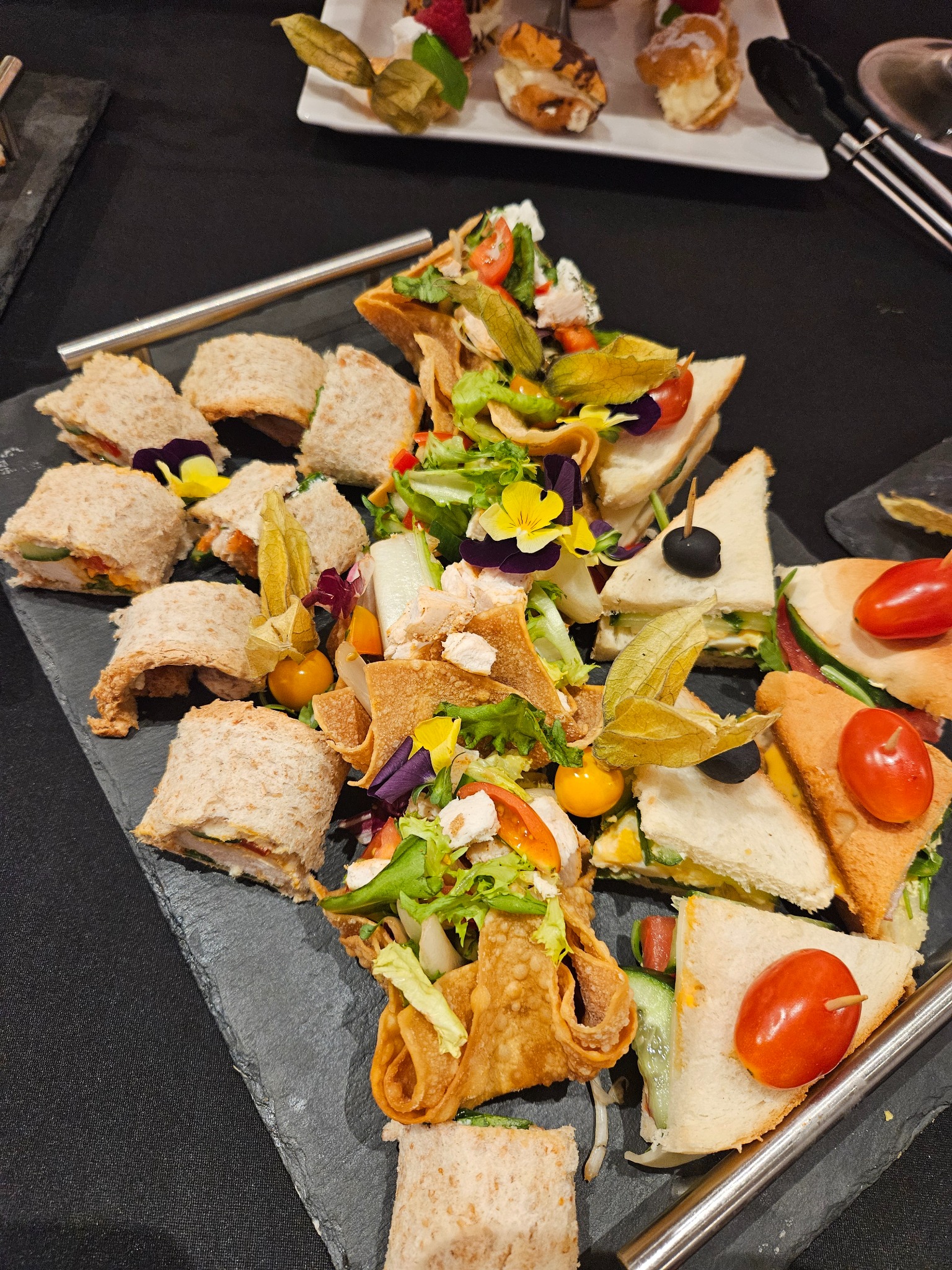 Elegancki catering: kanapki roladki, trójkątne, sałatki w jadalnych miseczkach, dekorowane kwiatami i miechunką, na czarnym kamiennym talerzu.