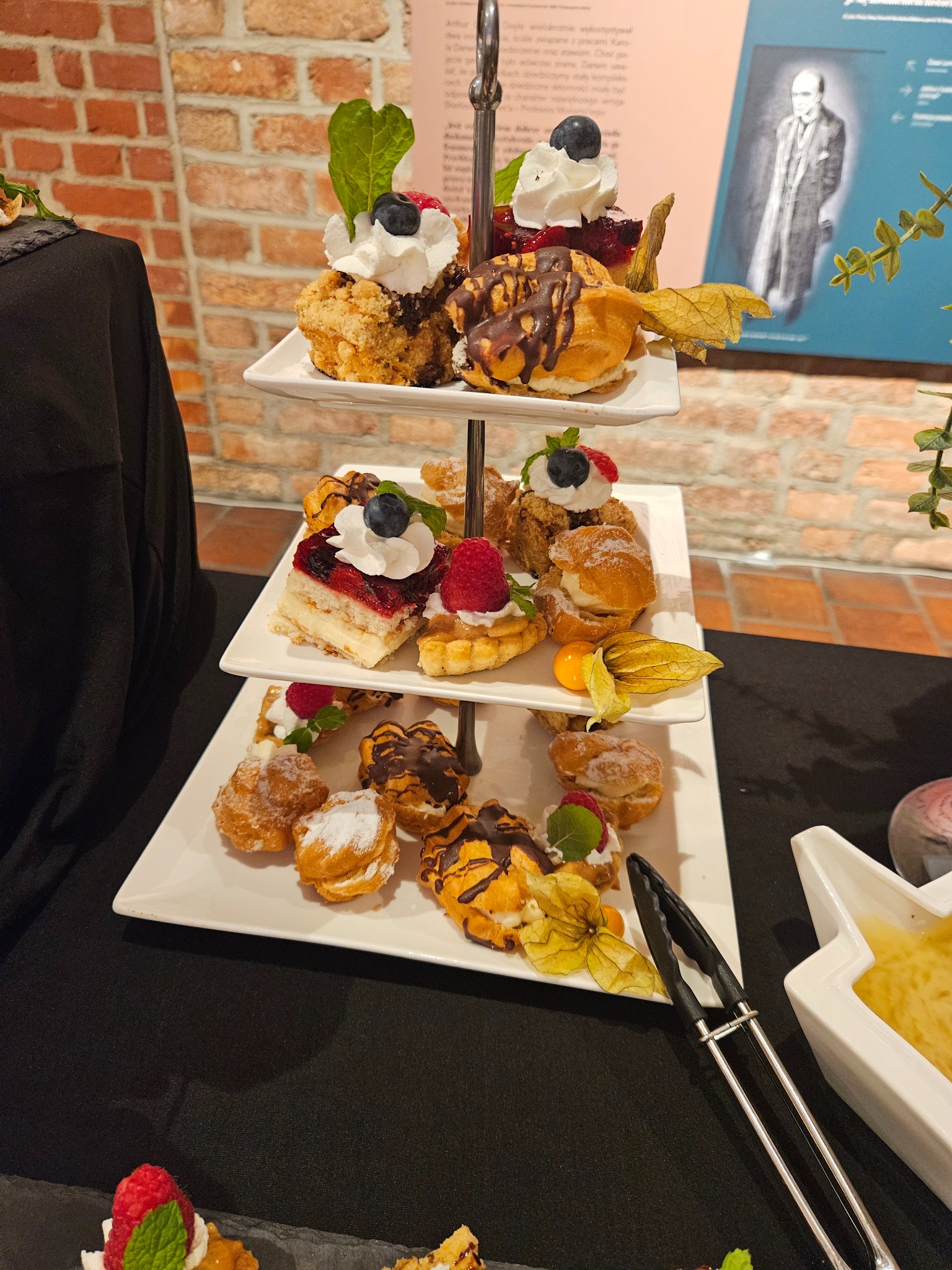 Trzypoziomowa patera z różnorodnymi, apetycznymi deserami: ciastka z kremem, tartaletki z owocami, eklerki z polewą, dekorowane physalis, na tle ceglanej ściany.